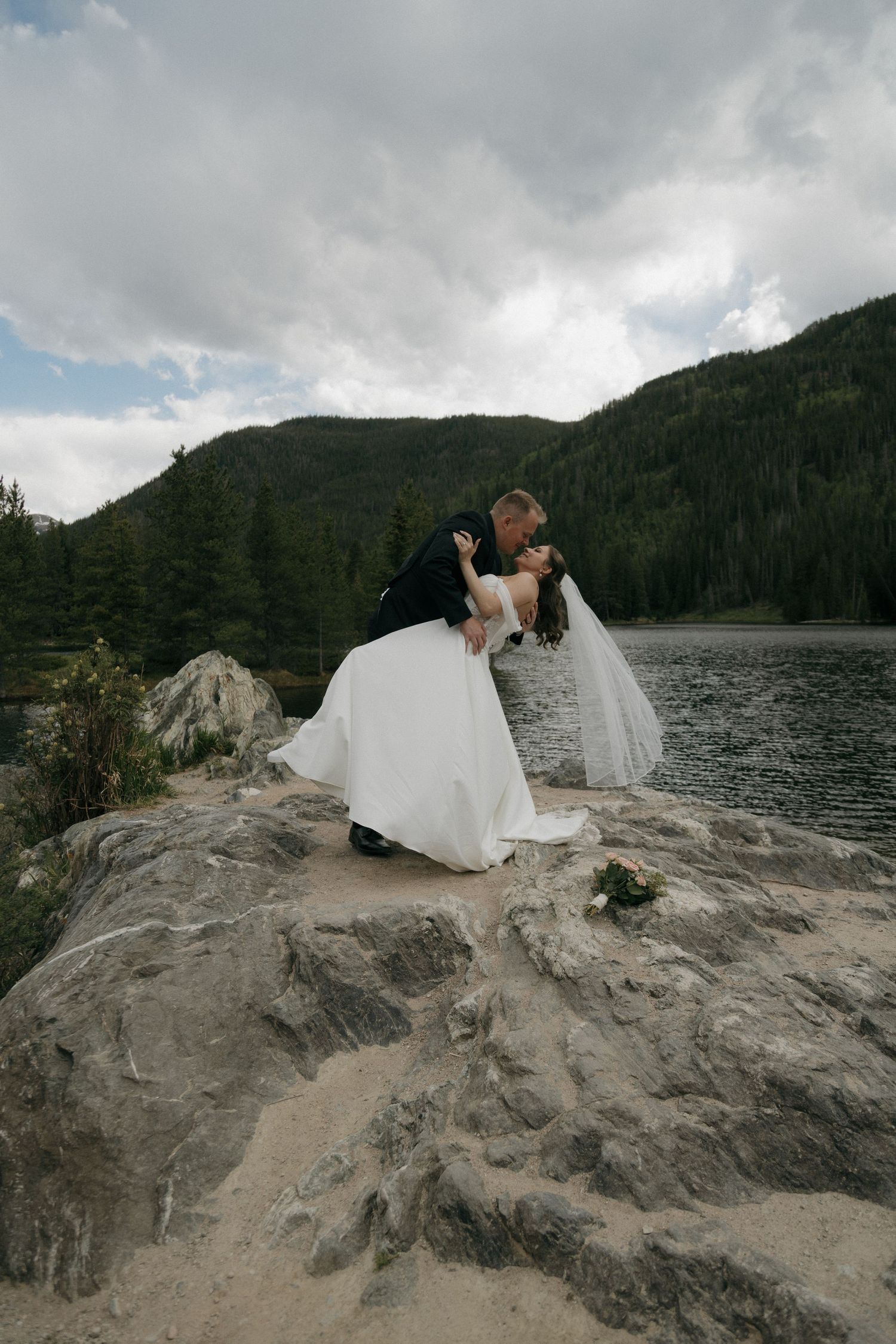 Groom dipping bride in a romantic kiss on rocky lakeside with mountain backdrop.