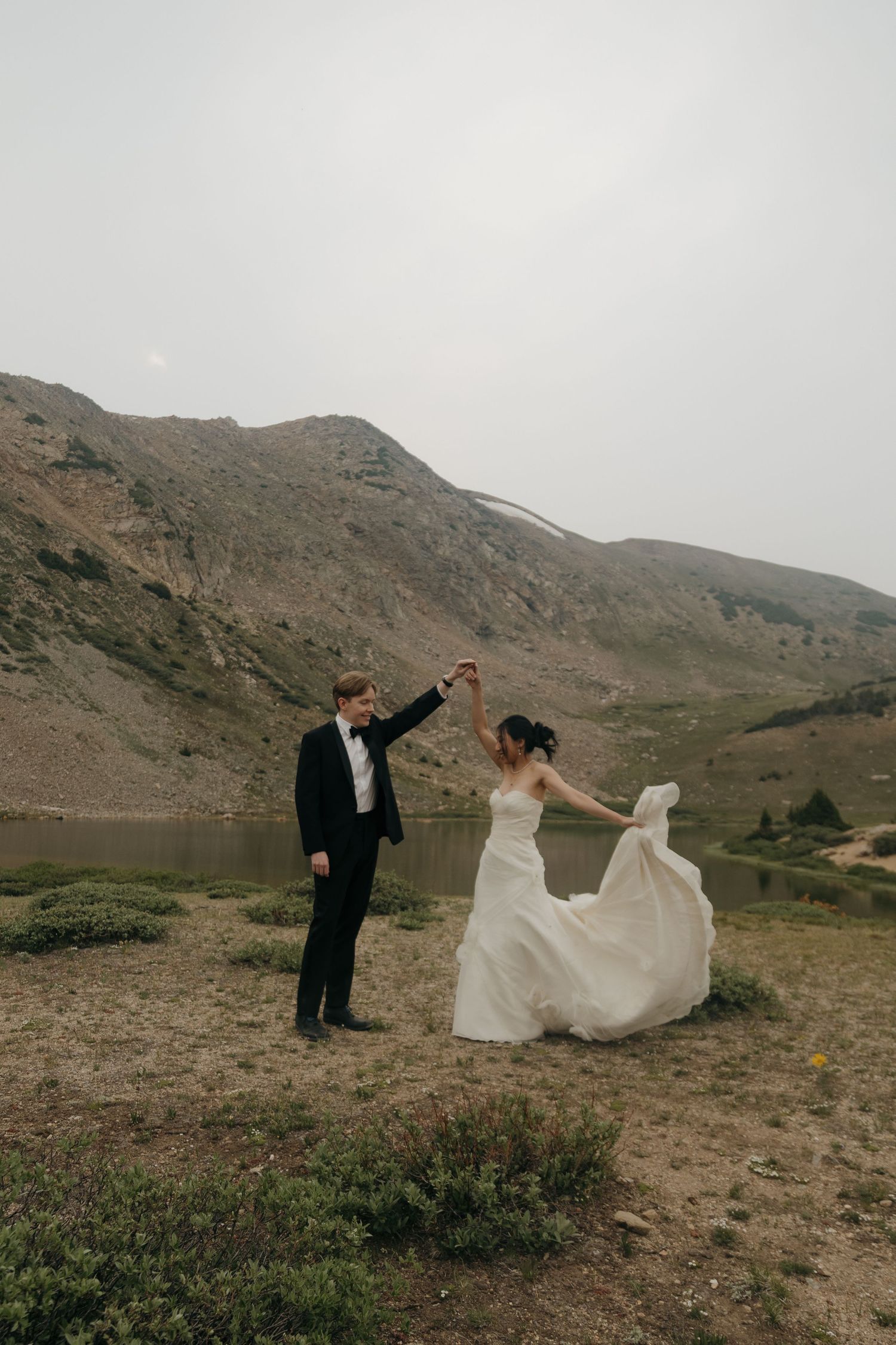 Bride and groom dancing by a mountain lake, her white gown flowing in the wind during an outdoor elopement.