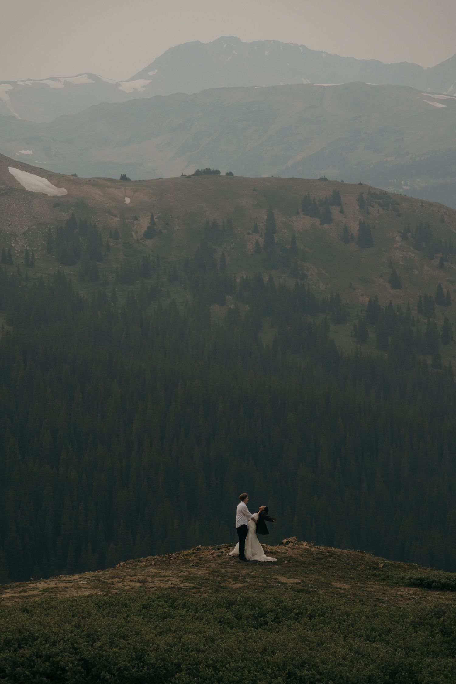 Bride and groom standing on a scenic mountain ridge with vast forested slopes and hazy peaks behind.