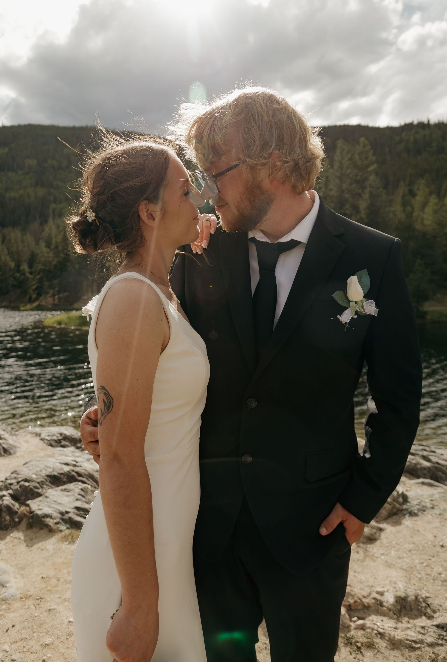 A newlywed couple gazes at each other lovingly outdoors, groom in dark suit with boutonniere, bride with tattoo.