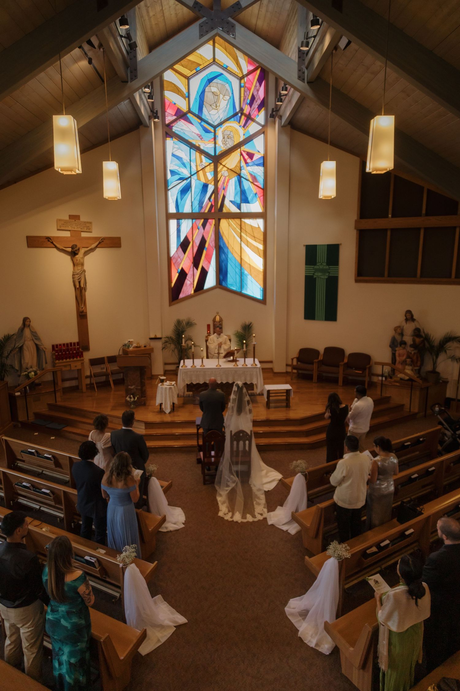Aerial view of intimate Catholic wedding ceremony with colorful stained glass window above altar.