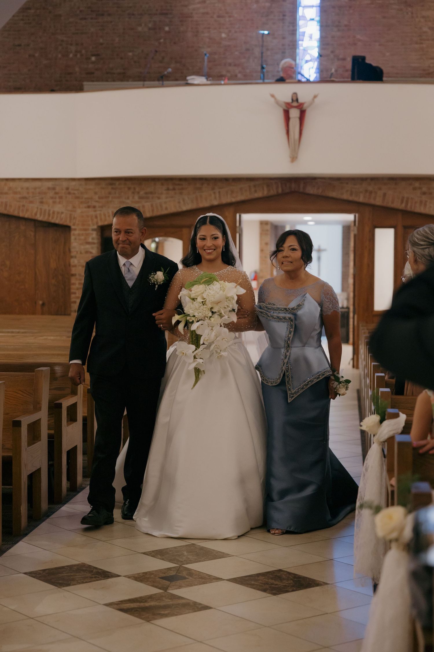 Bride walks down church aisle escorted by parents, wearing white ball gown and holding white floral bouquet.
