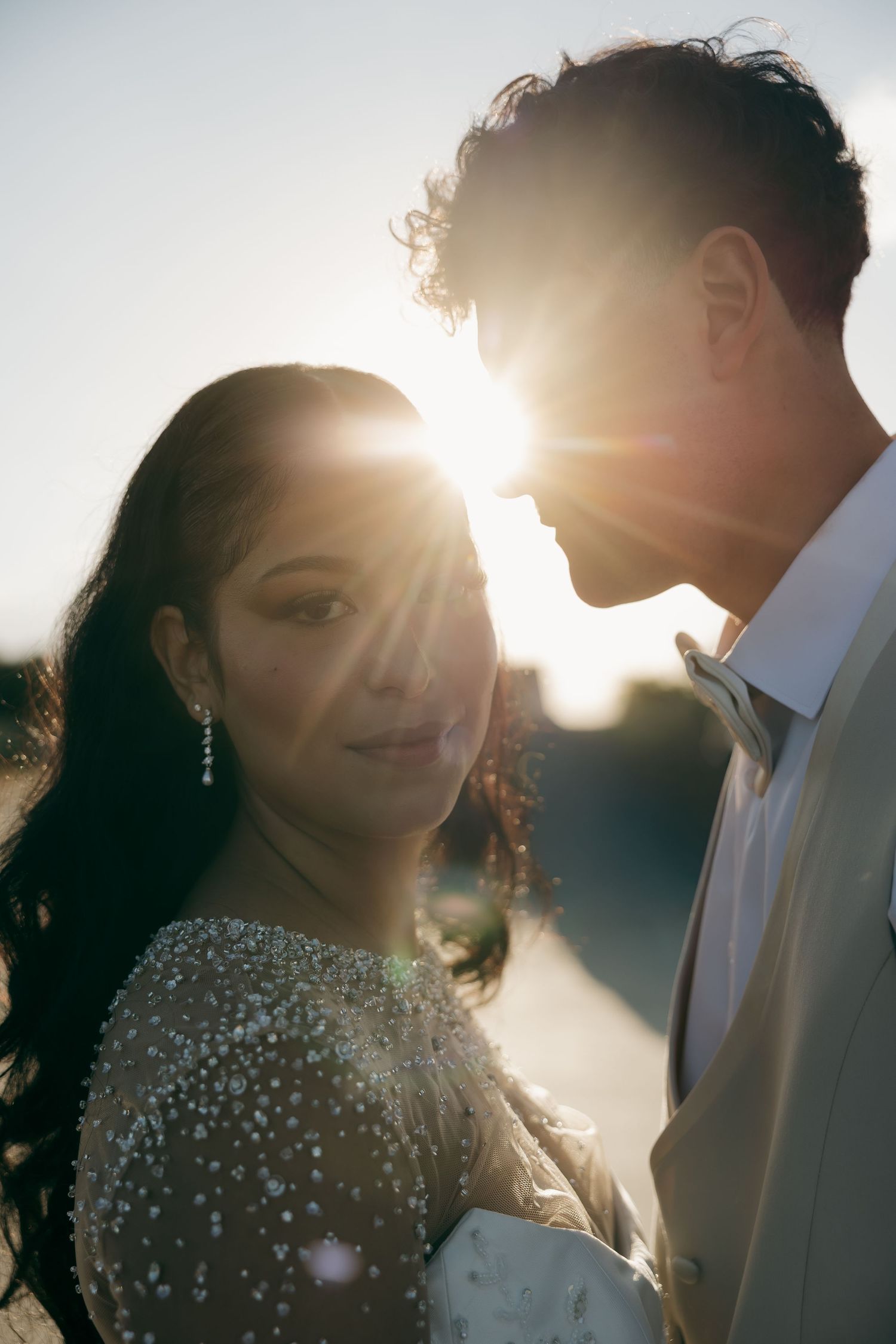 Couple in wedding attire share a romantic moment with golden sunset backlight creating a stunning lens flare effect.