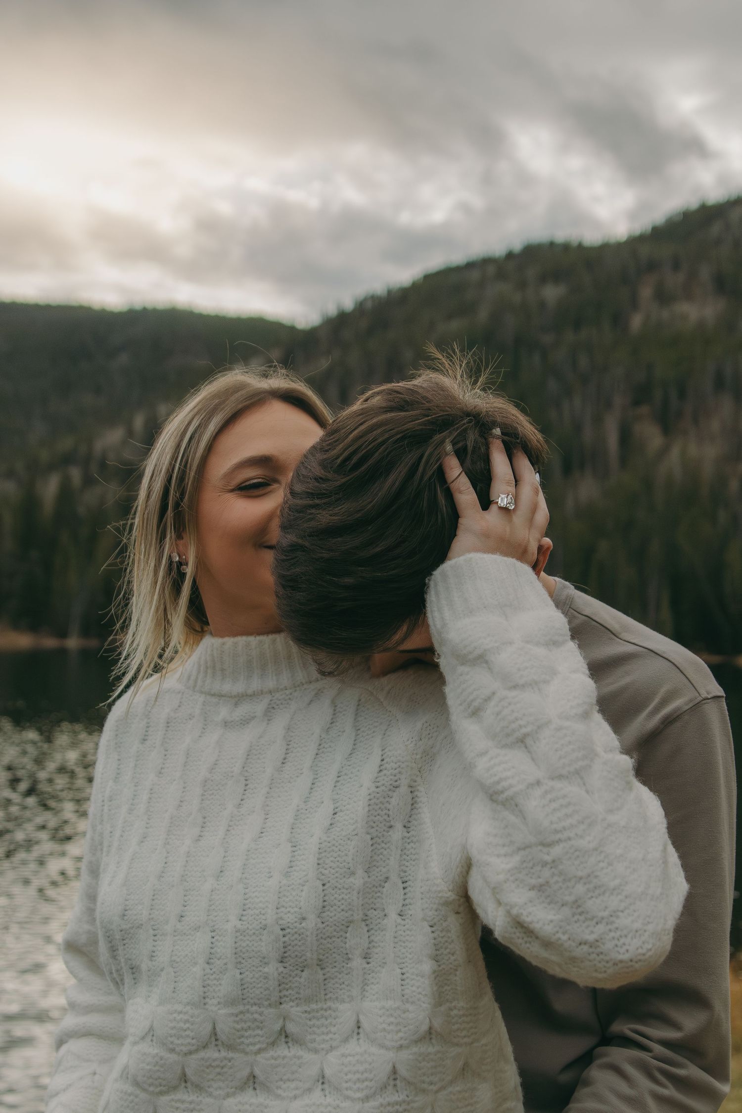 Two people in cozy sweaters share an intimate moment by a mountain lake on an overcast winter day.