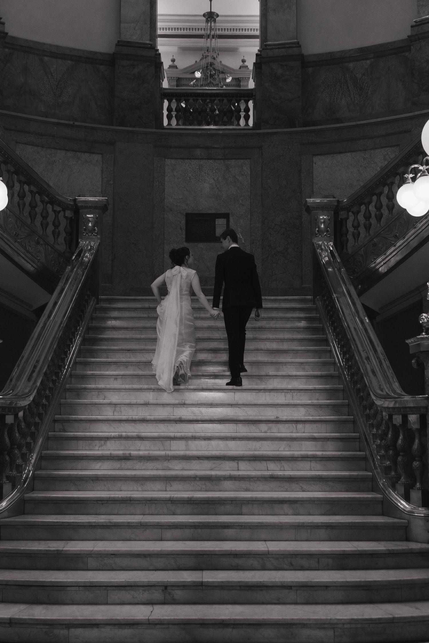 Two figures ascend an elegant grand staircase in a formal building, captured in black and white photography.