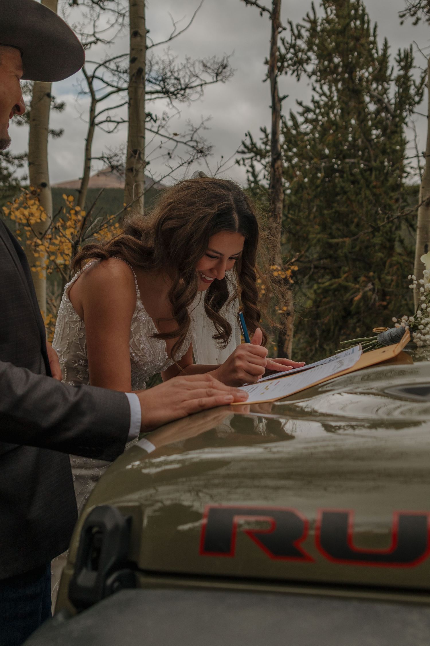 A couple signs documents on a Jeep Rubicon hood in a mountain forest setting.
