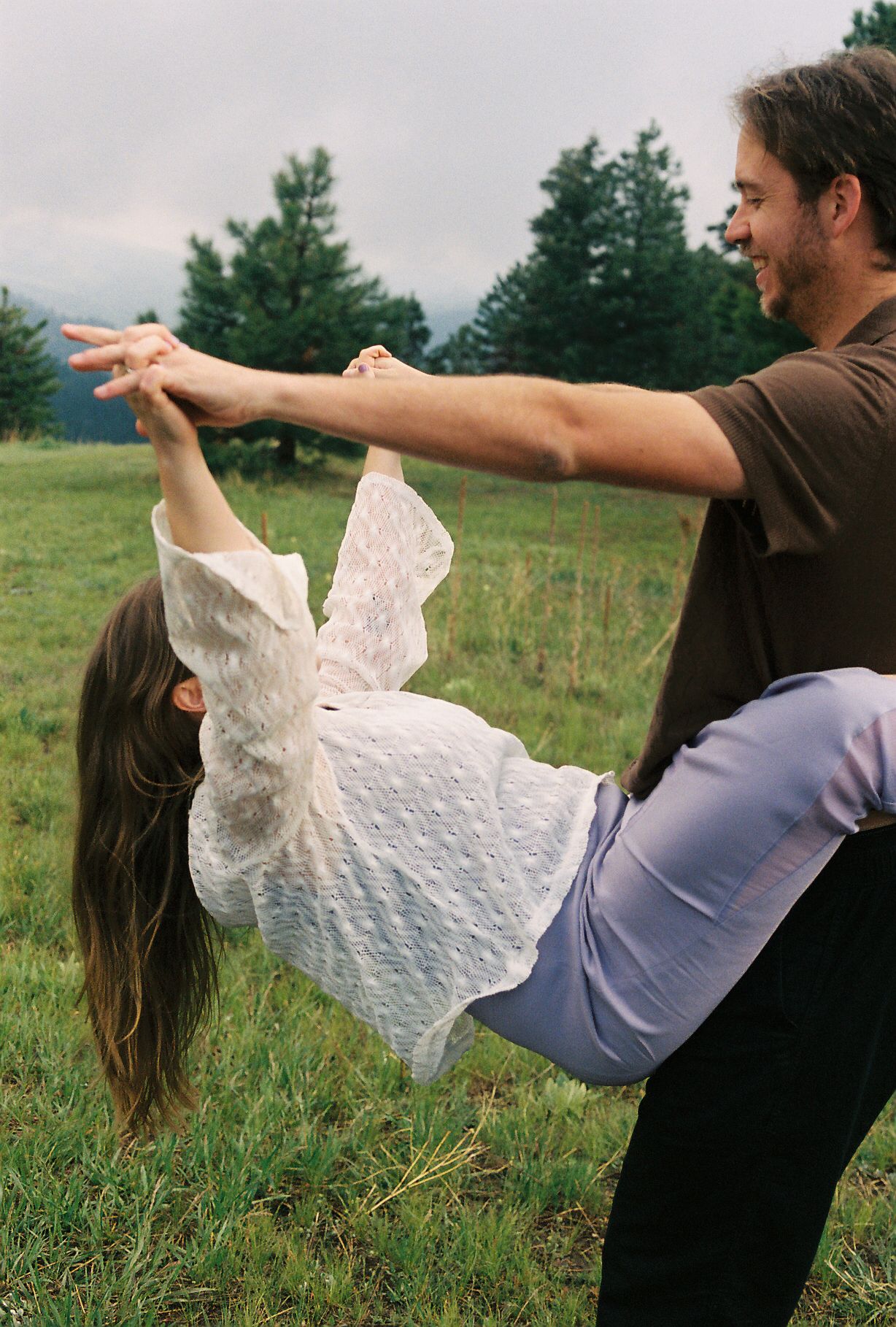 Man playfully dipping woman in a white lace top on a green grassy field with trees in background.