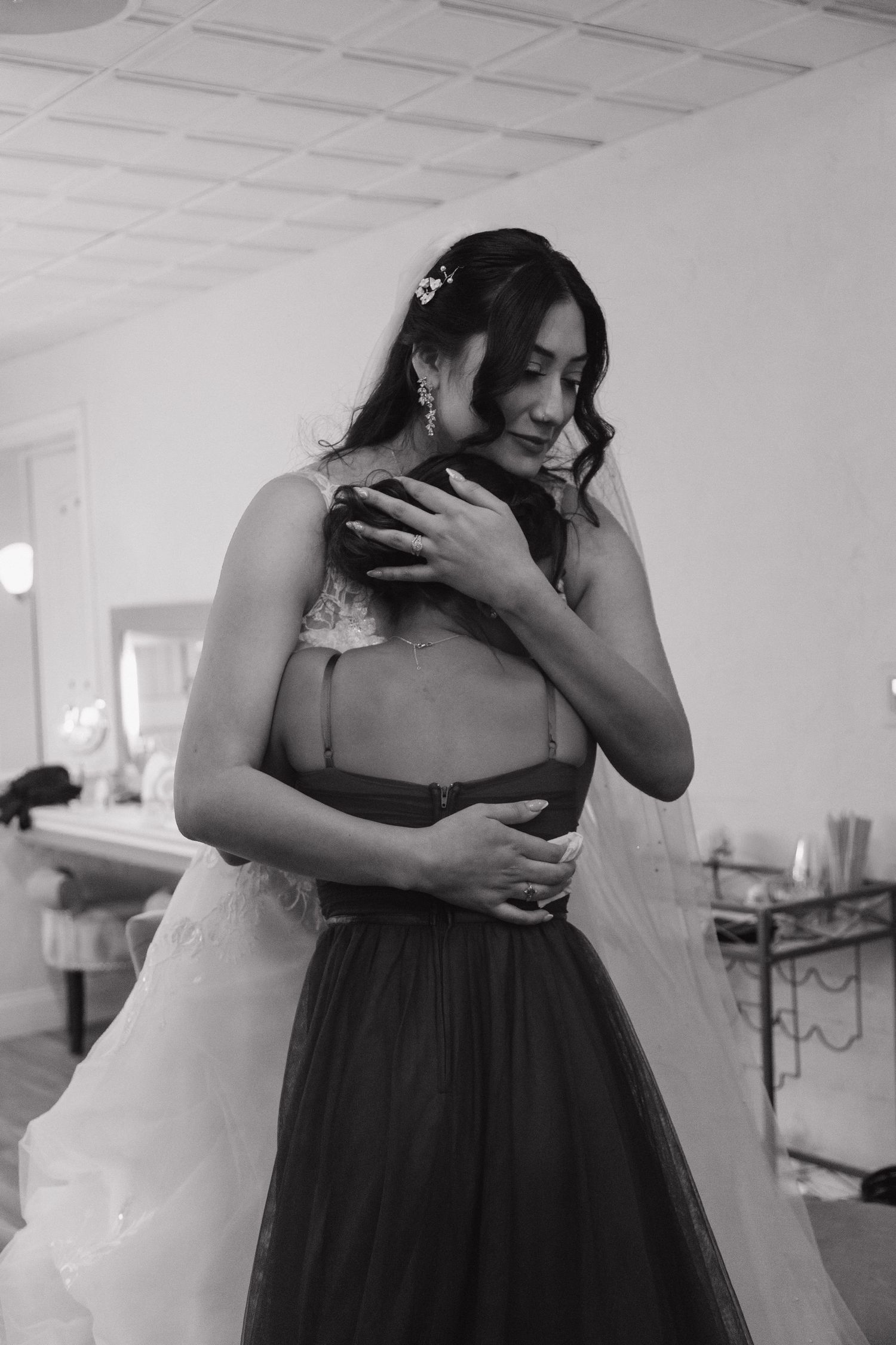 Emotional bride in white gown shares tender embrace with bridesmaid in black dress before wedding.