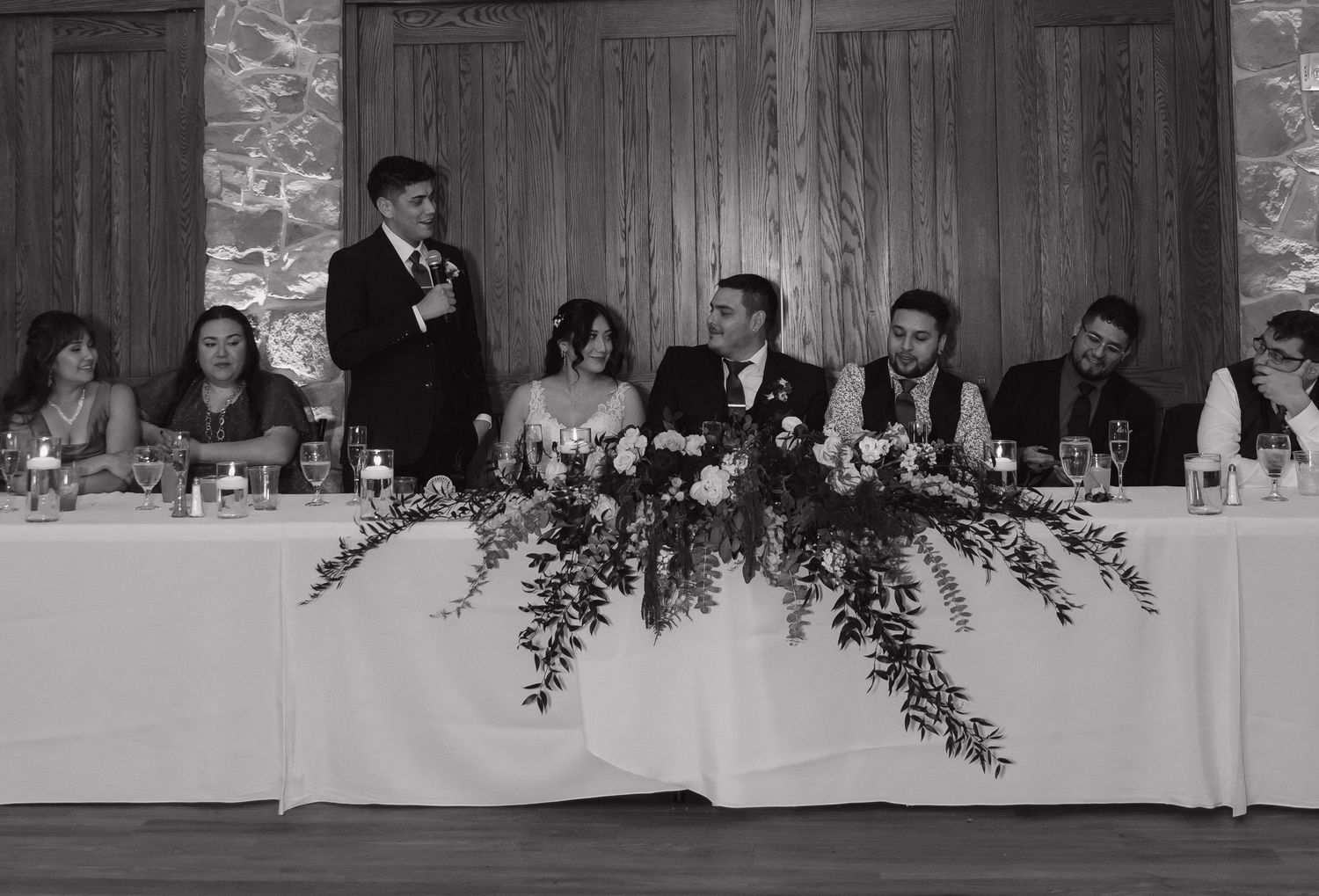 Black and white photo of wedding party at head table during reception toast speech.