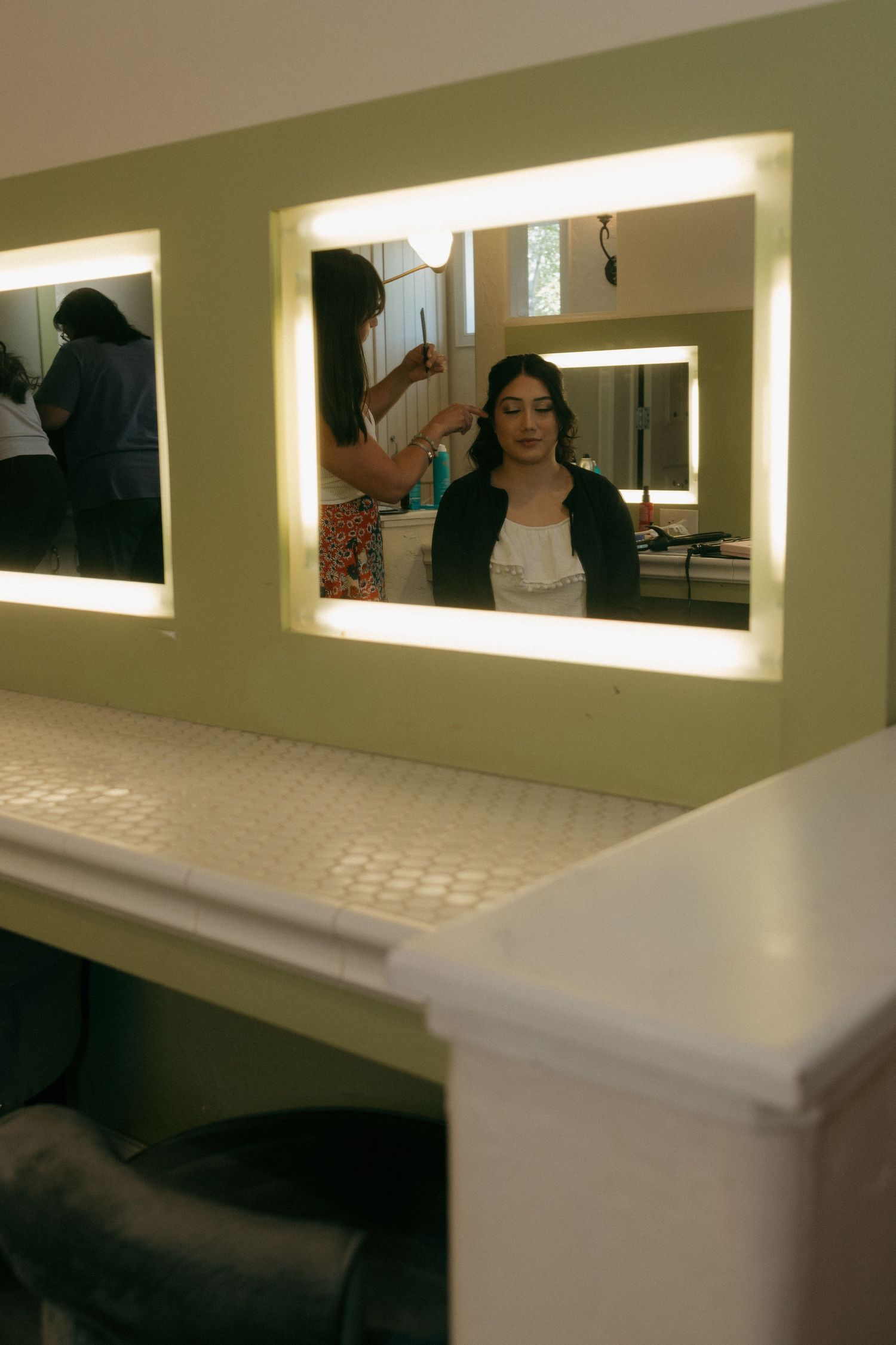Bride getting hair styled reflected in a lighted vanity mirror in a bridal preparation room.