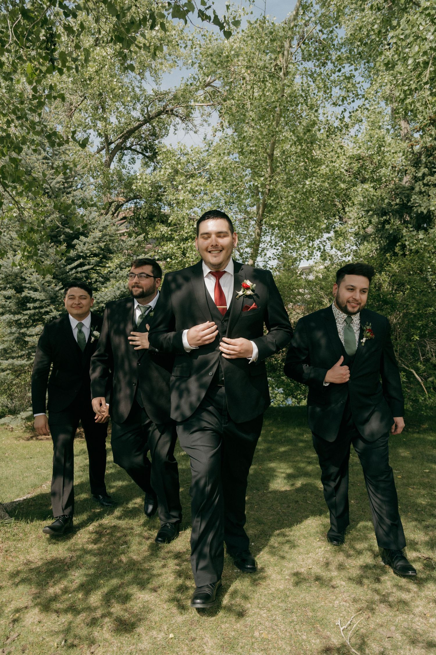 Smiling groom in black suit with red tie leads groomsmen walking outdoors among green trees.