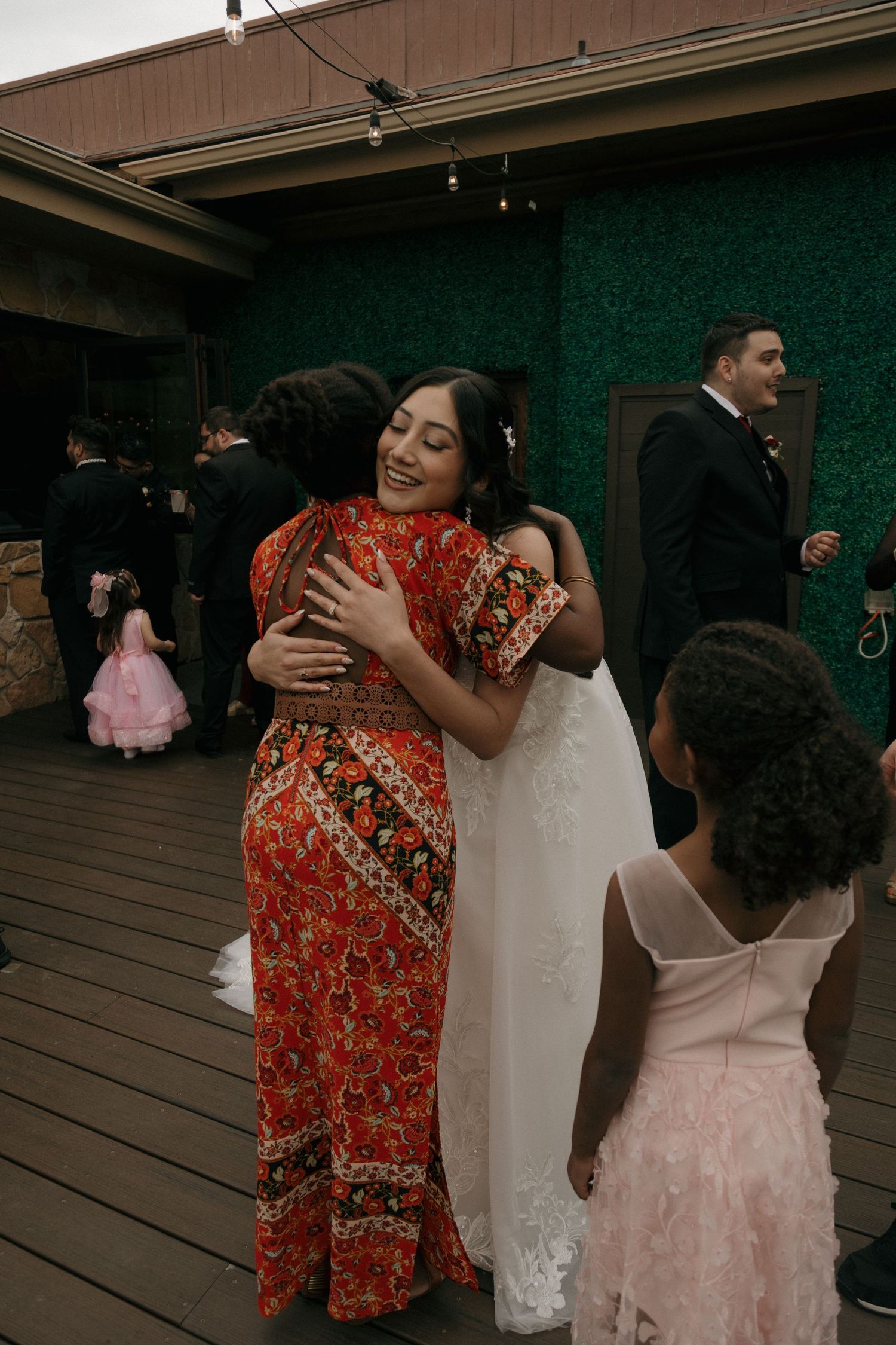 Smiling bride in white lace gown shares a warm hug with a guest in a vibrant red floral dress.