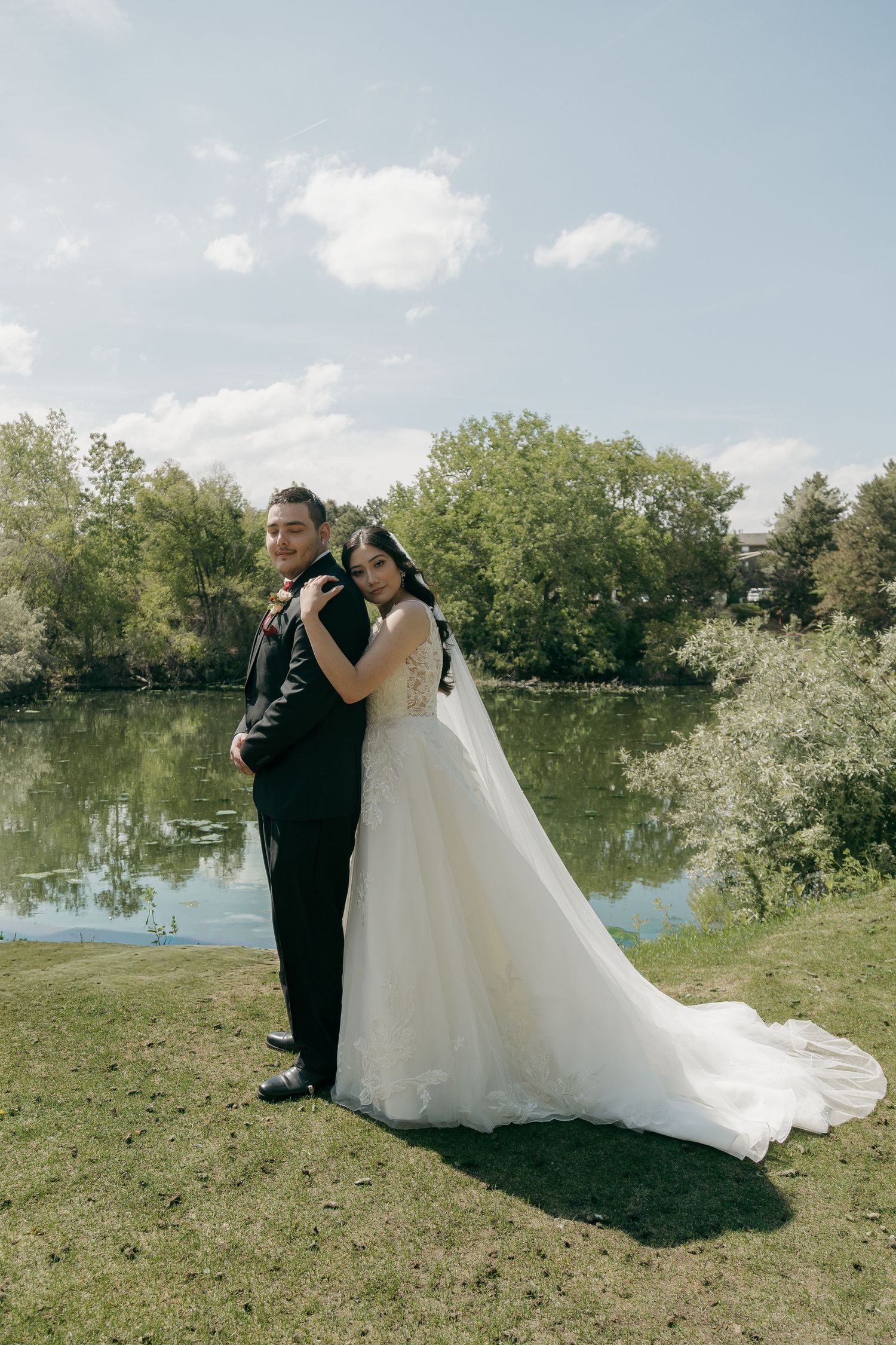 Bride and groom pose back-to-back in formal wedding attire beside a serene lake surrounded by lush green trees.