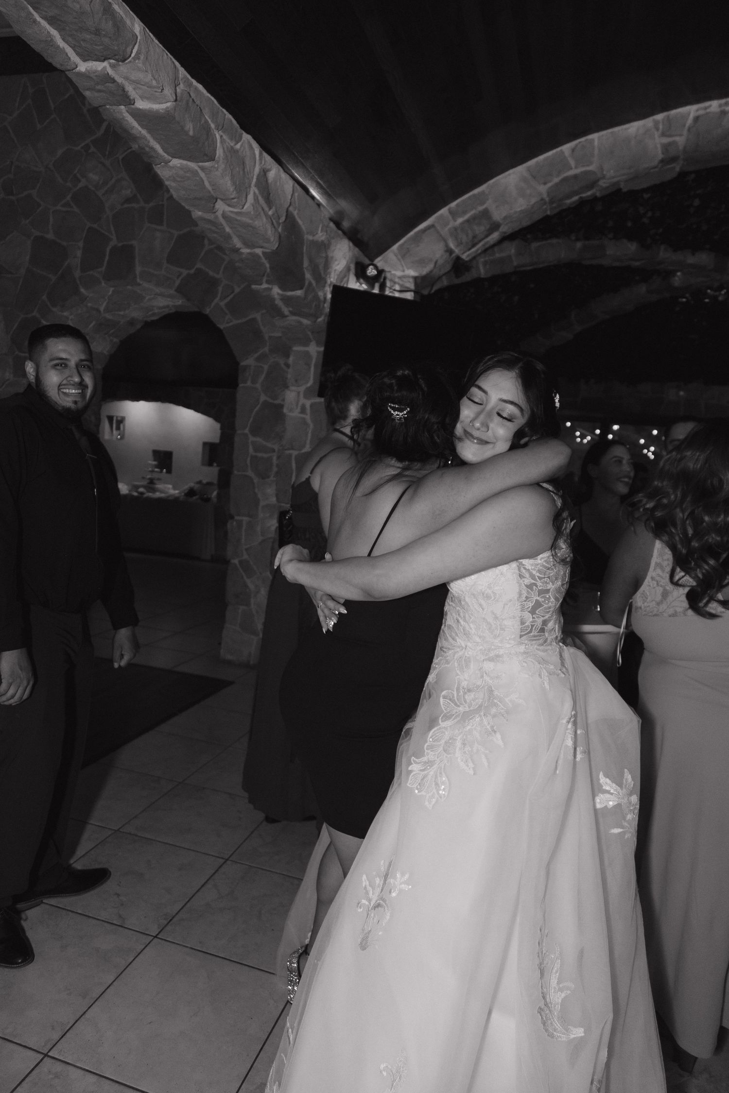 Black and white photo of bride in lace gown sharing a warm hug with a guest at her wedding.
