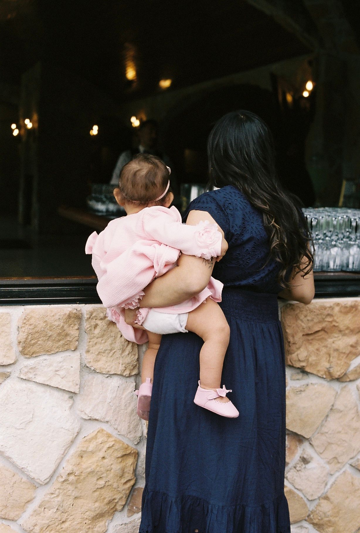 Mother in navy dress holds baby in pink outfit near stone wall at wedding venue.