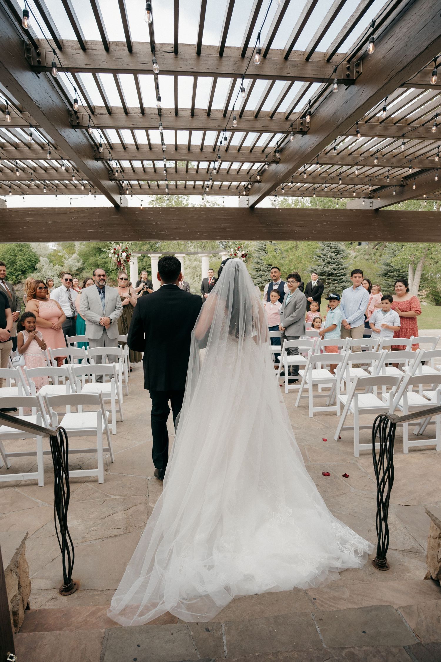 Bride and groom walk down the aisle under a wooden pergola with seated wedding guests applauding.