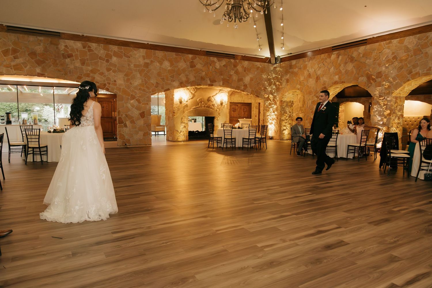 Bride and groom share first dance on hardwood floor in elegant stone-walled wedding reception ballroom.