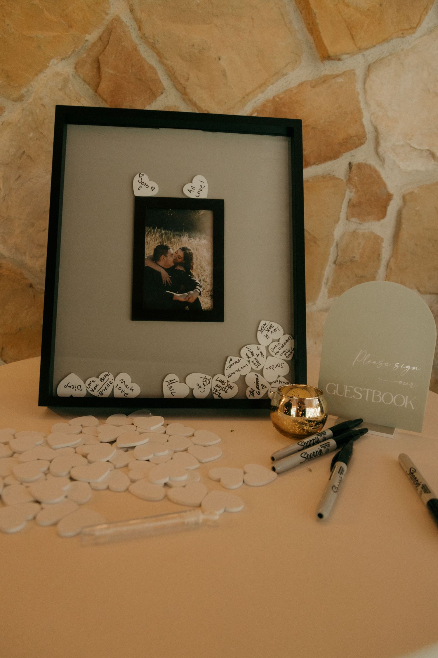 Wedding guestbook shadow box frame with heart-shaped signatures, couple photo, markers, and decorative gold bowl.