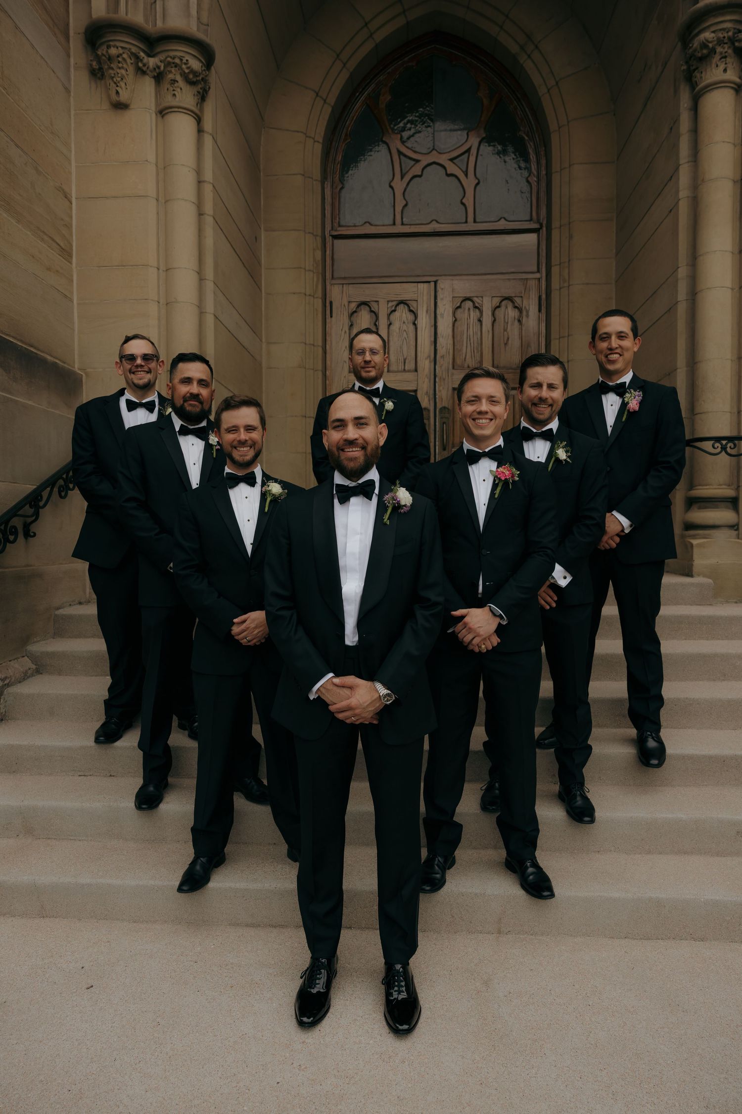 Groom and groomsmen in black tuxedos with bow ties pose on stone church steps for a formal wedding photo.