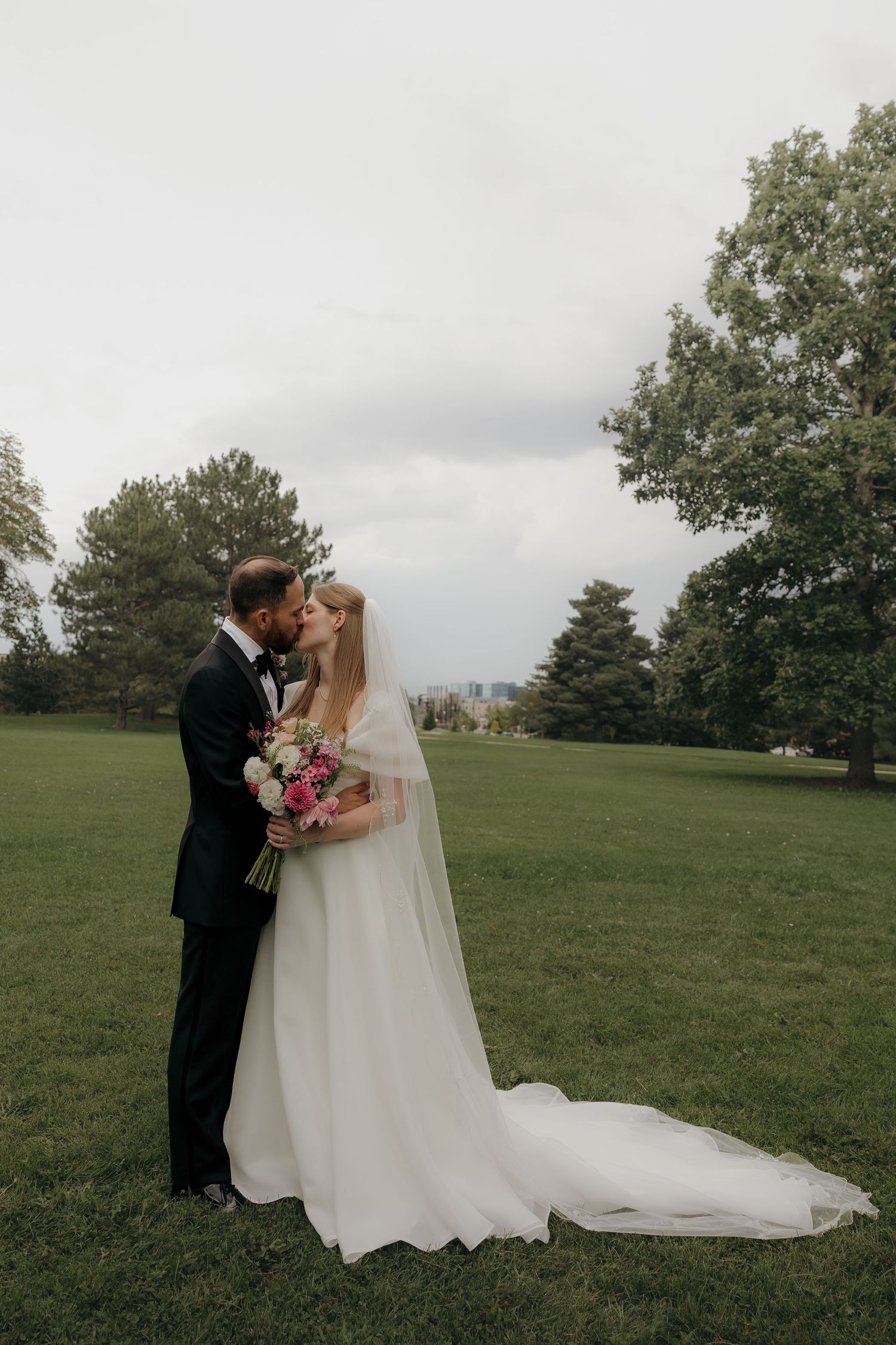 Bride and groom share romantic kiss in green park setting, bride holding pink floral bouquet.
