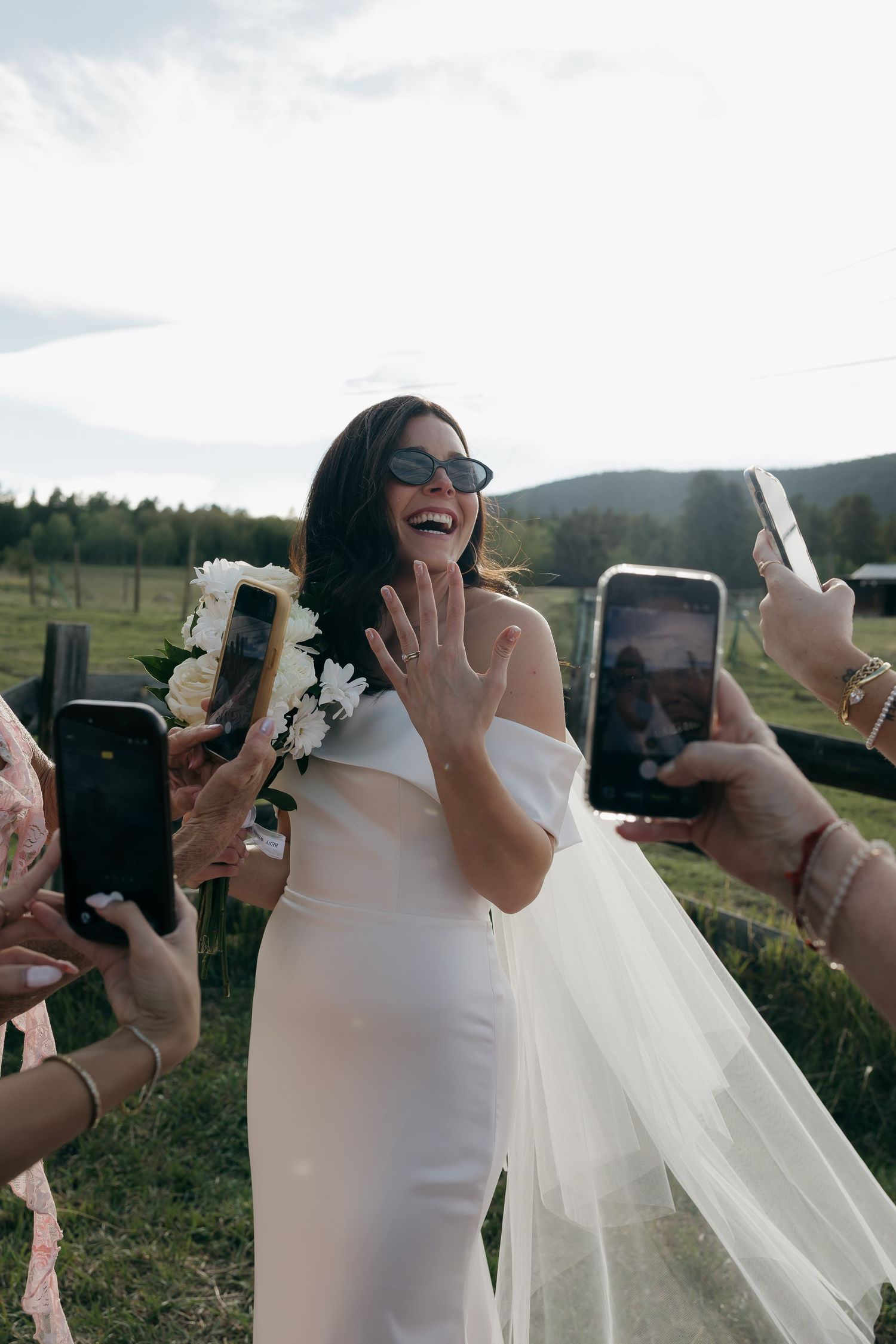 Radiant bride in white gown showing ring while guests photograph her outdoors.