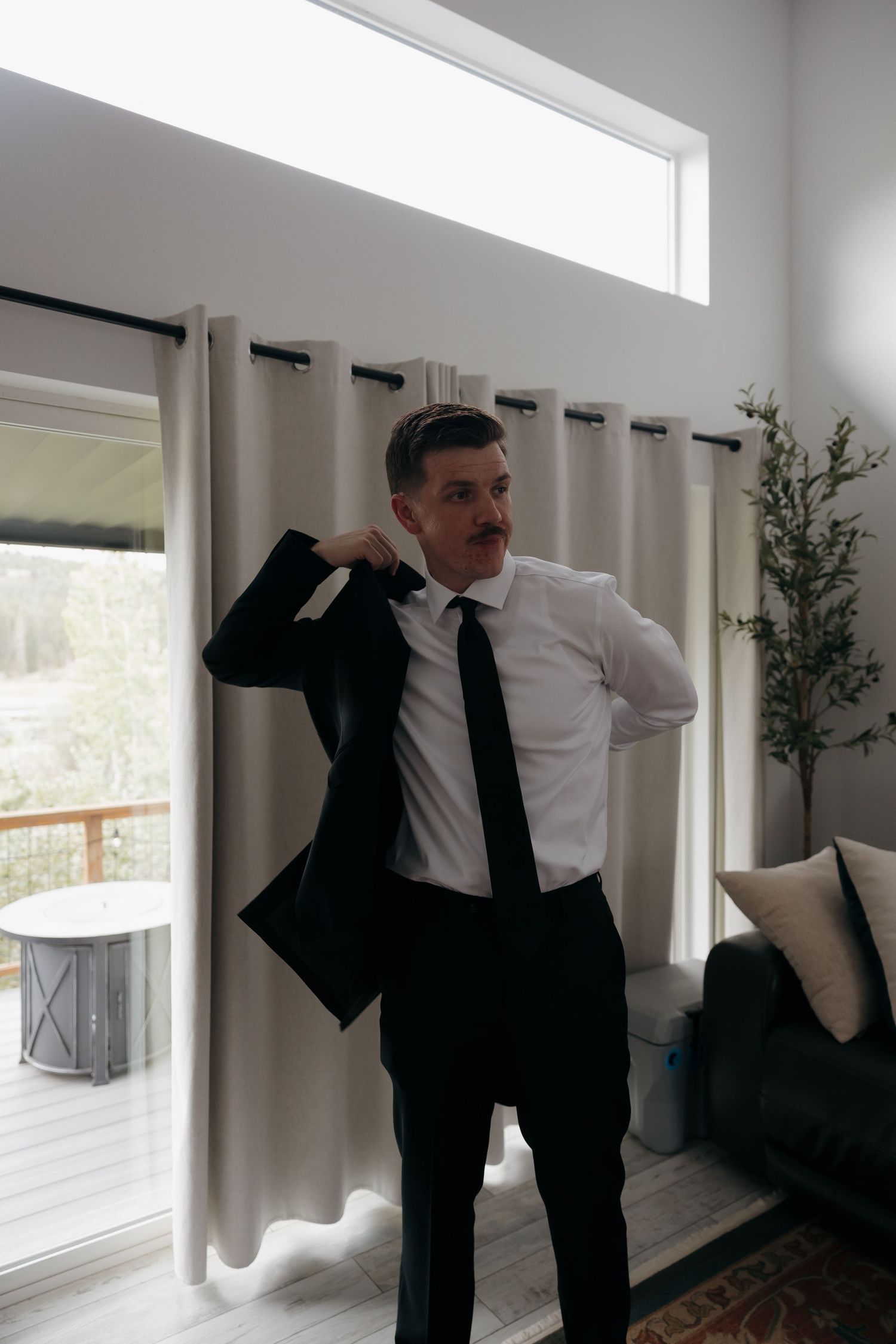Groom in black suit and tie putting on jacket indoors before wedding ceremony.