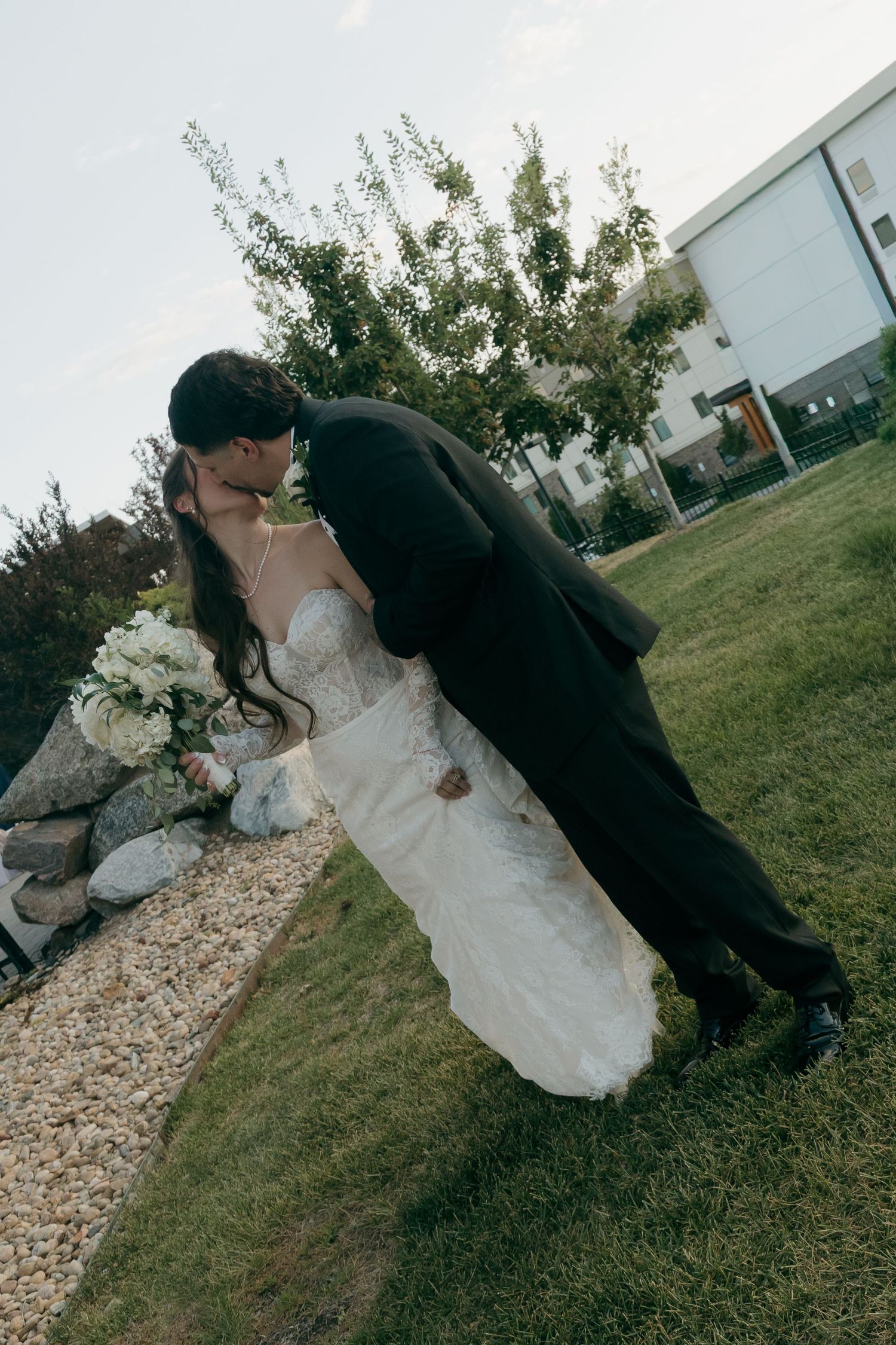 Groom dipping bride for a romantic kiss outdoors on green lawn in wedding attire.