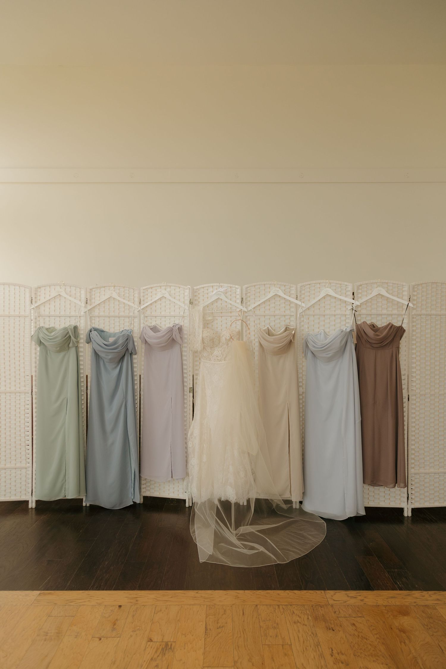 Bridesmaid dresses in pastel colors hanging on white room divider with bridal gown and veil.