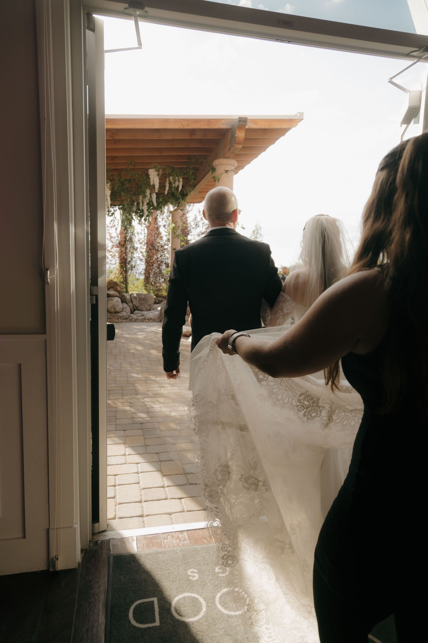 Bride in lace gown with veil being carried walks through doorway with groom at outdoor wedding venue.