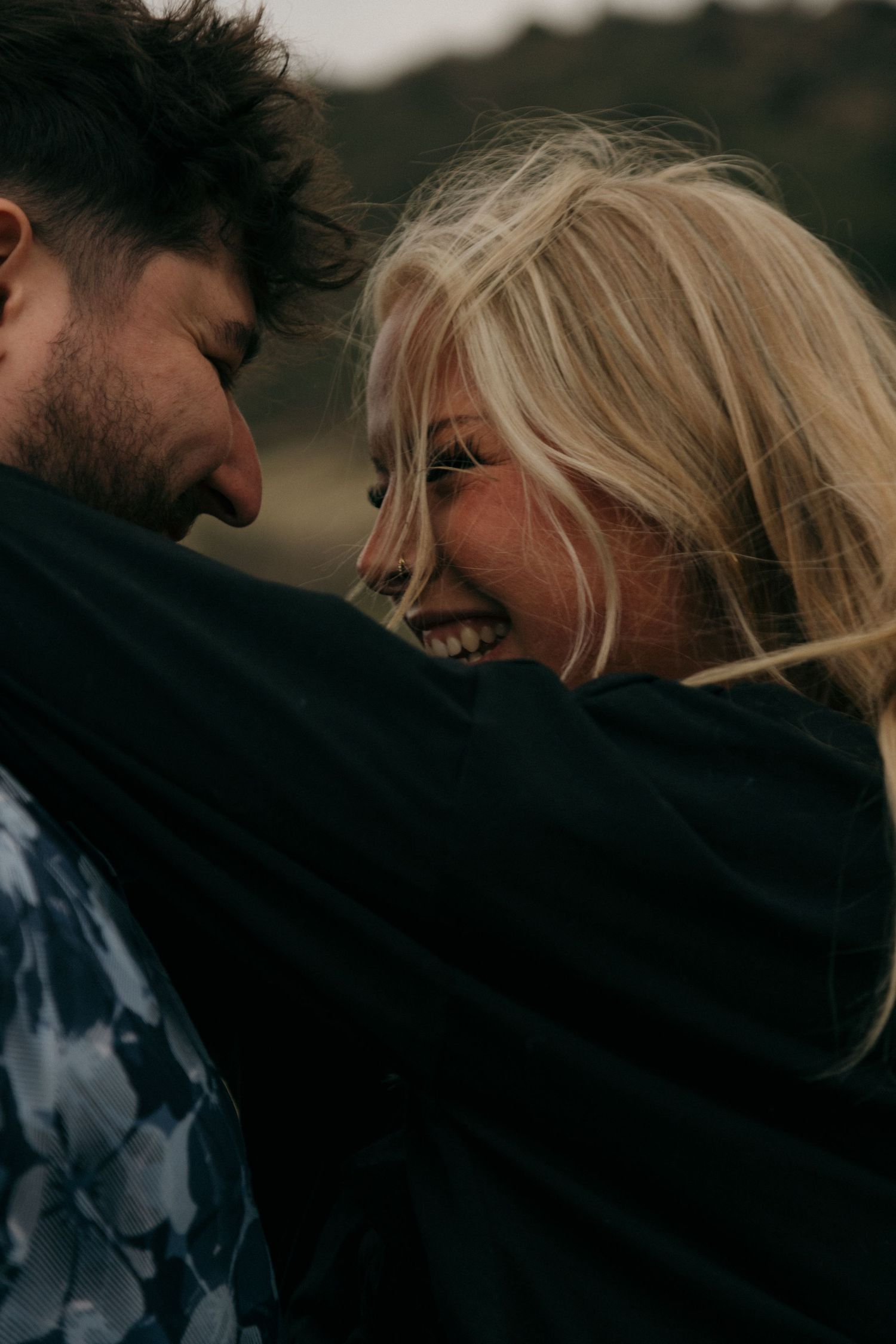 A laughing blonde woman and dark-haired man share a joyful intimate moment outdoors in a moody natural setting.