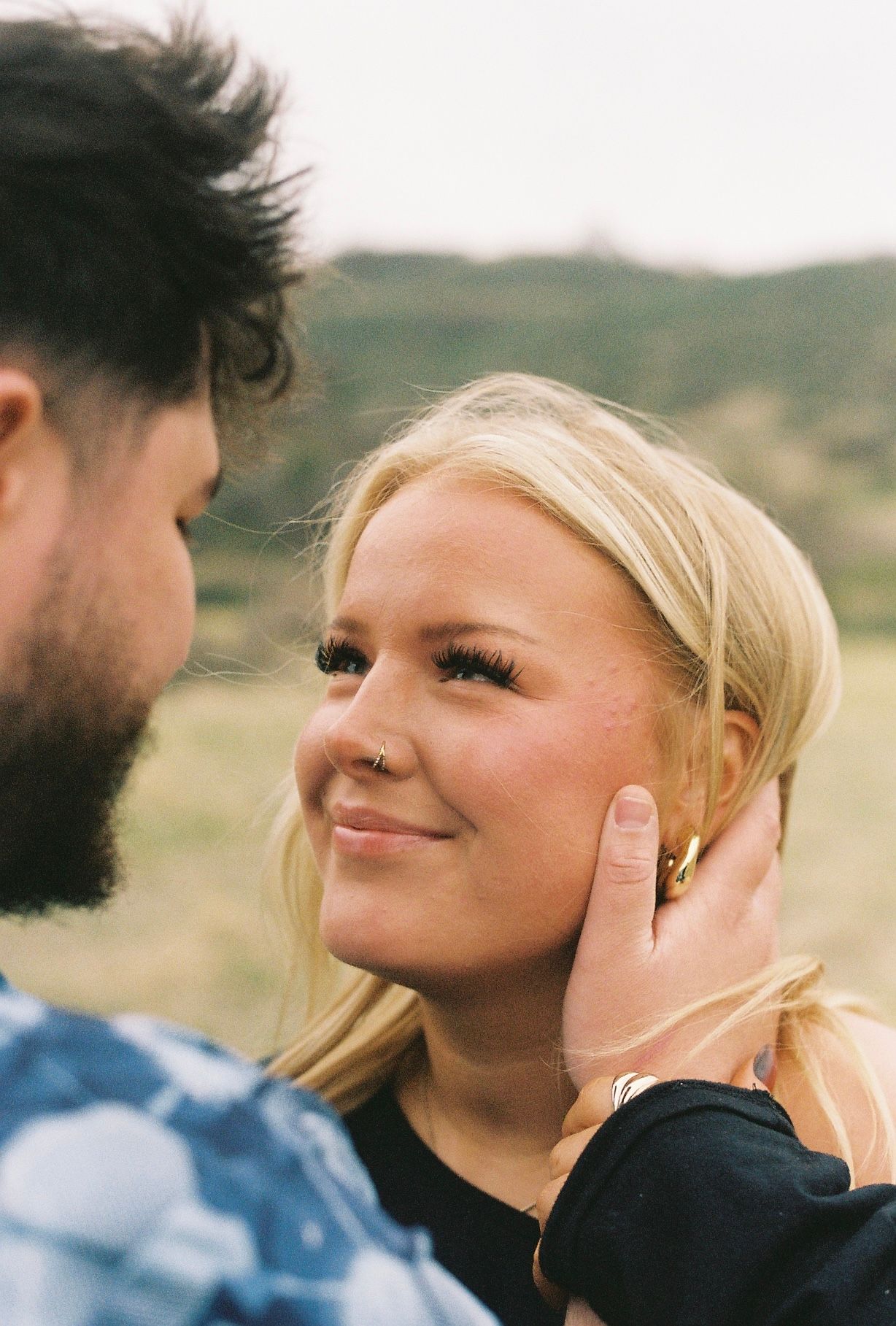 A smiling blonde woman with a nose ring looks up at a bearded man touching her face outdoors.