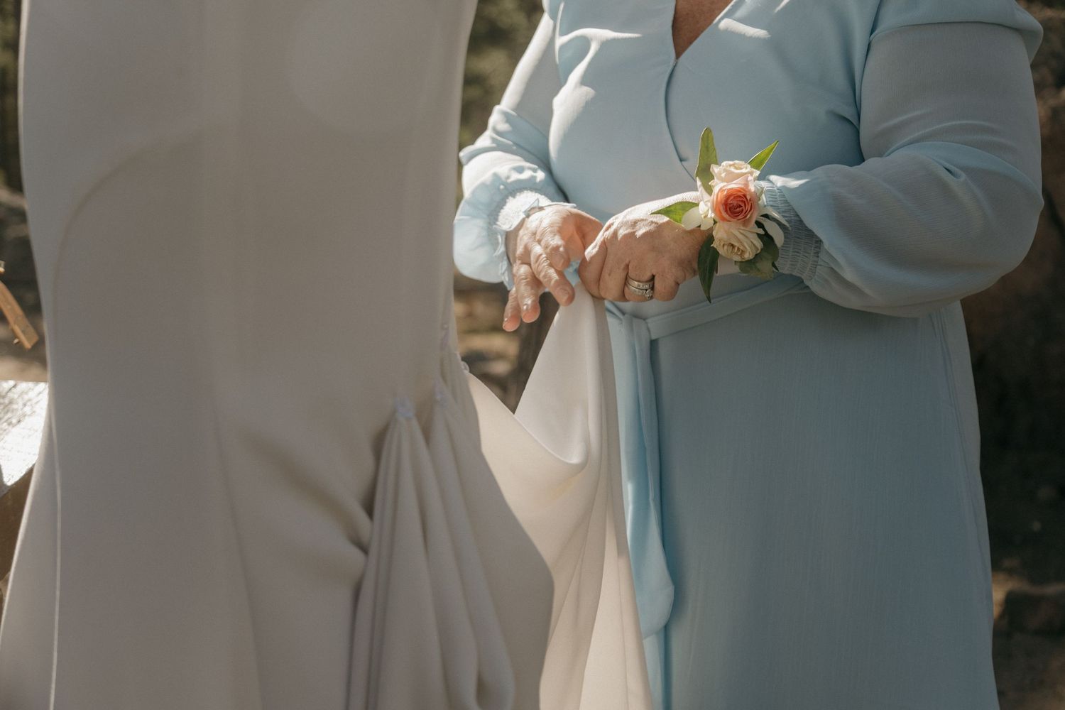 Woman in light blue dress with floral corsage holding bride's hand at outdoor wedding.