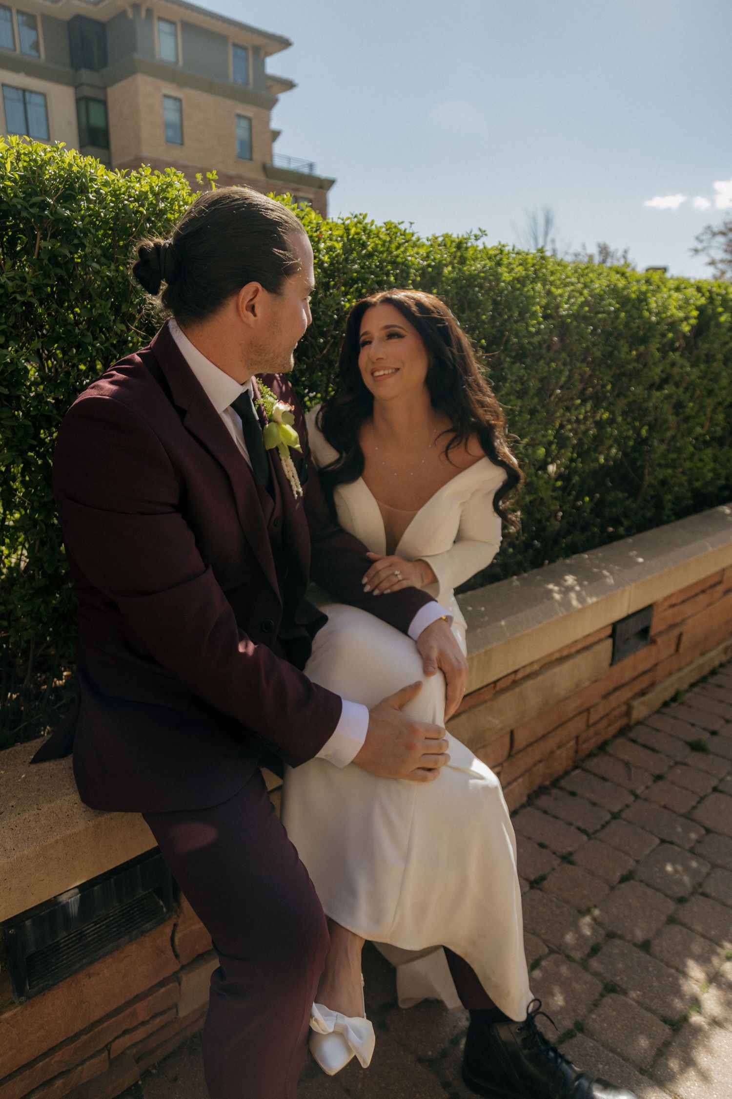 Bride and groom sitting together on stone ledge surrounded by lush green hedges on wedding day.