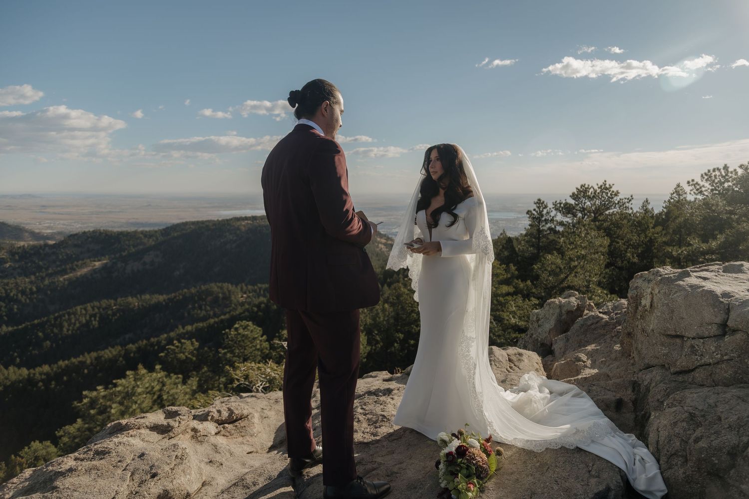 A bride and groom stand on a rocky mountain summit exchanging vows under a vast blue sky.