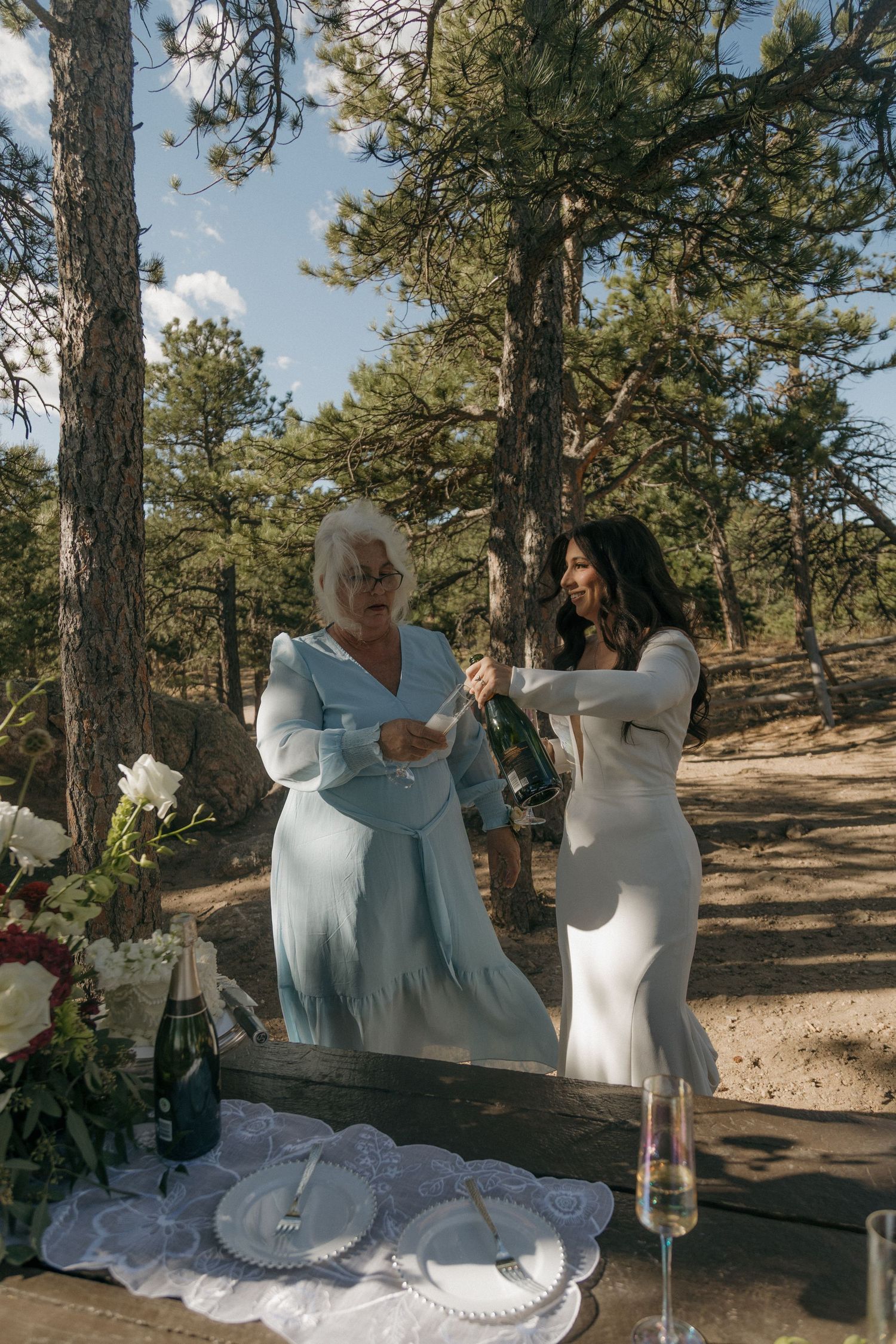 Bride in white dress and older woman in blue opening a champagne bottle at an outdoor wedding celebration.