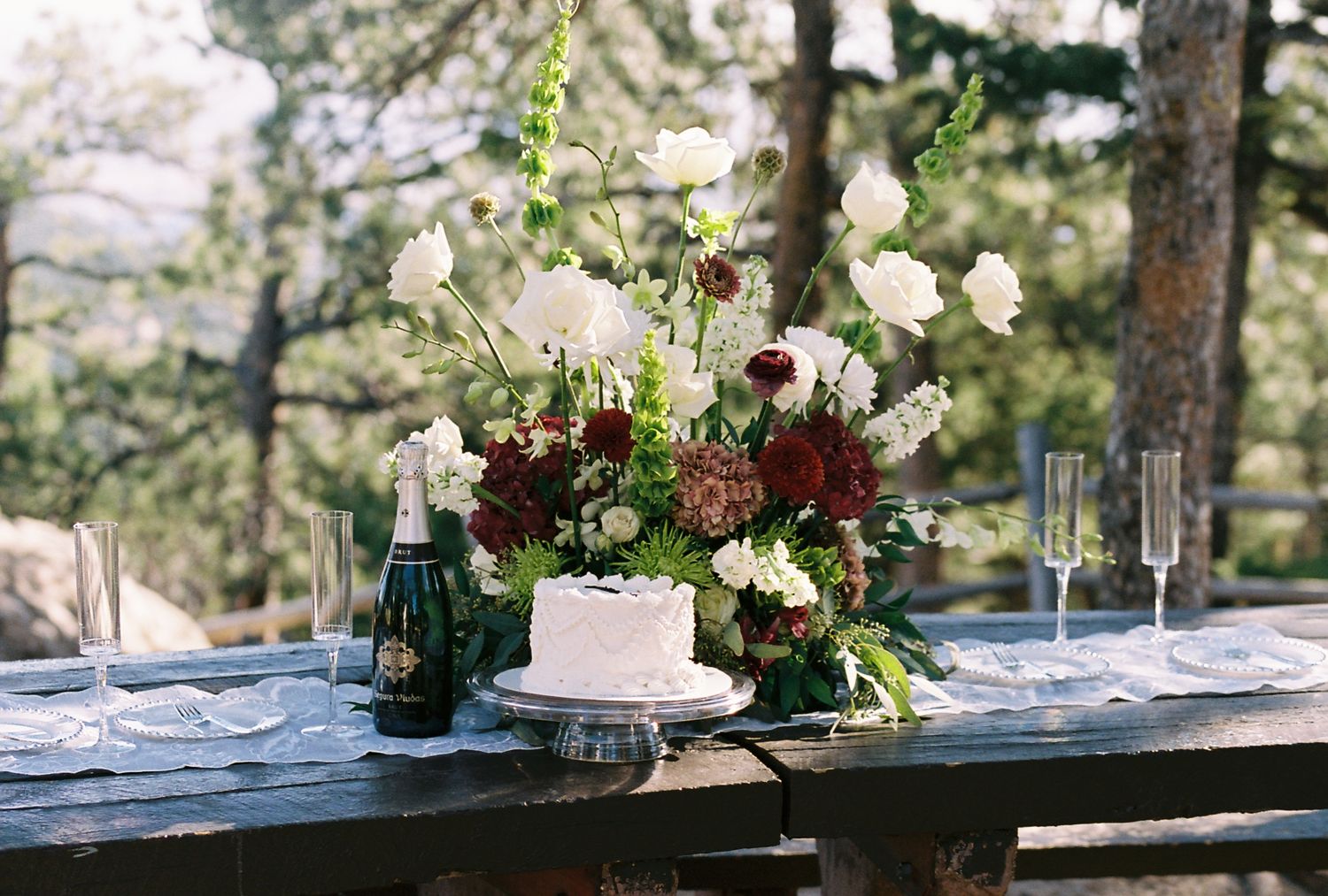 Elegant outdoor wedding cake table with lush white and burgundy floral arrangement and champagne bottle.