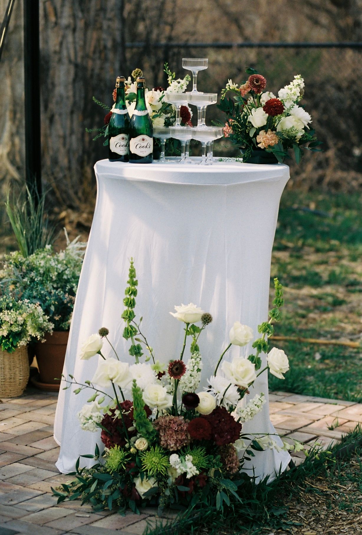 Elegant white cocktail table with champagne tower and white and burgundy floral arrangements at outdoor wedding.
