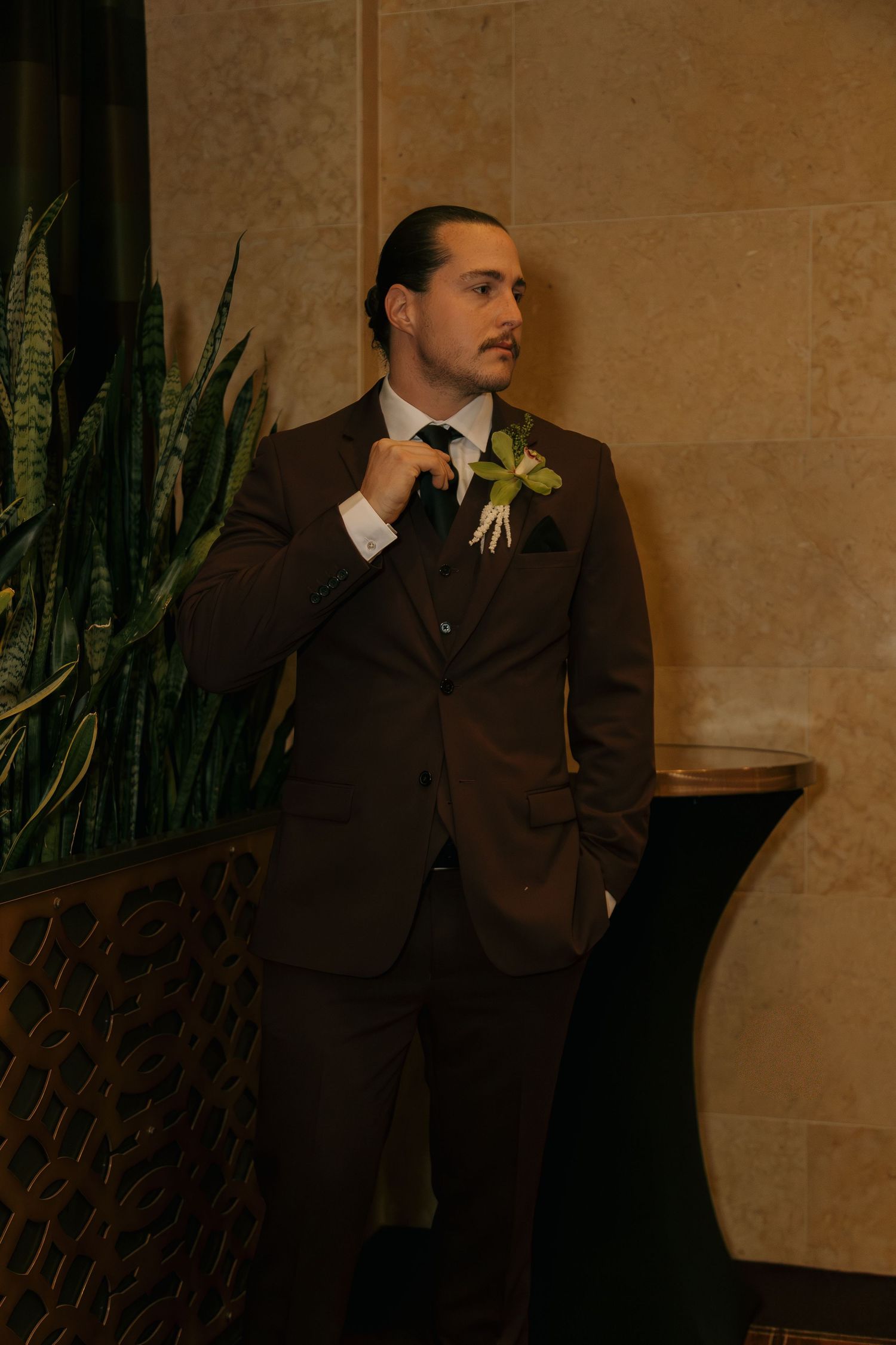 Groom in brown suit with floral boutonniere standing near indoor plants.