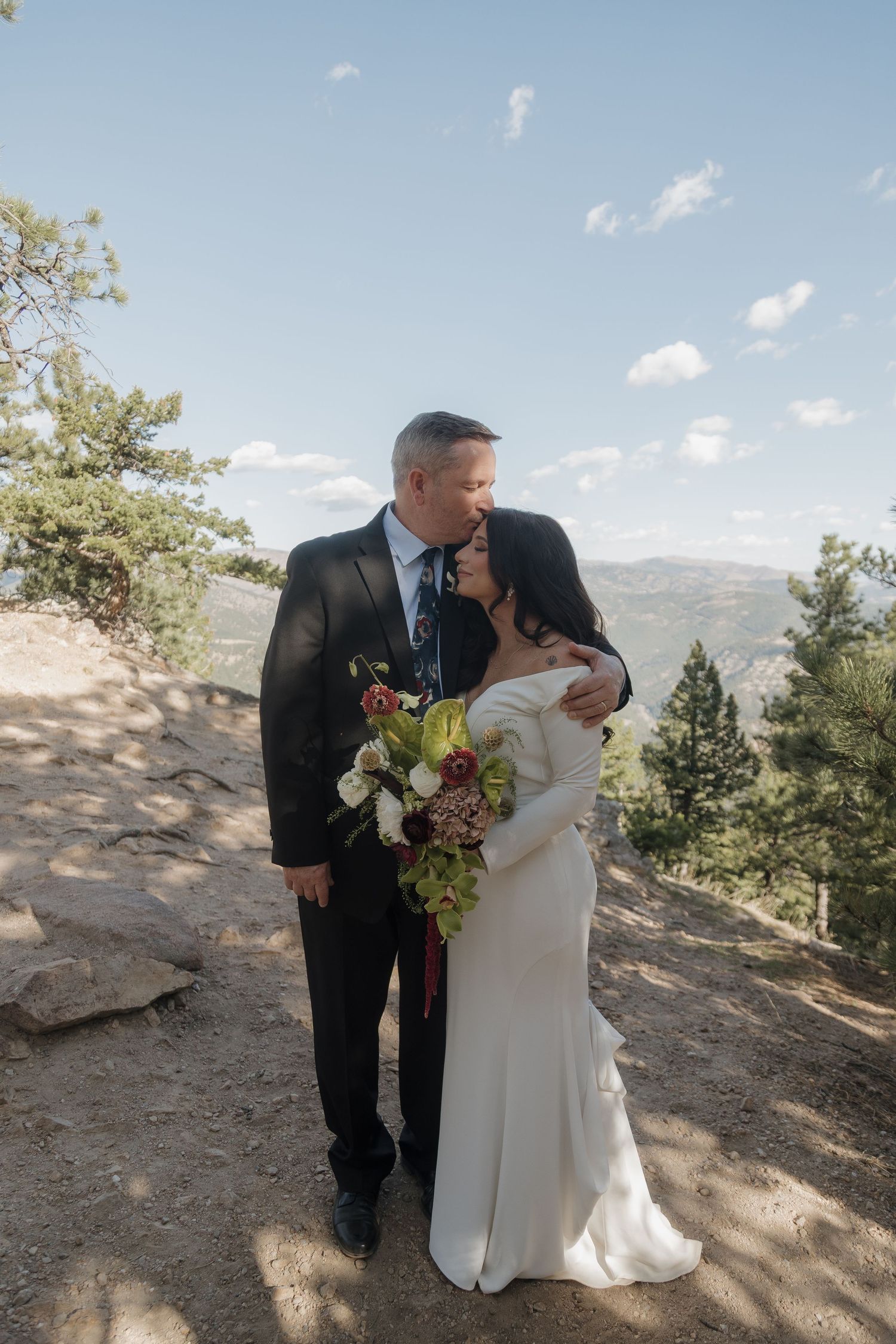 Groom in black suit kisses bride on forehead while she holds a burgundy and white bridal bouquet outdoors.