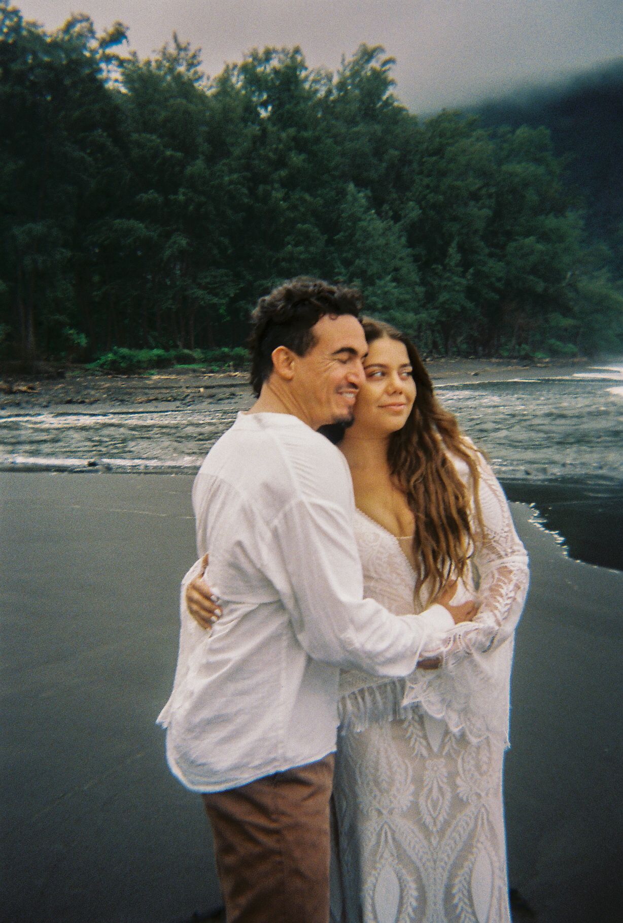 Couple embraces on black sand beach with lush green trees and misty mountains behind them.