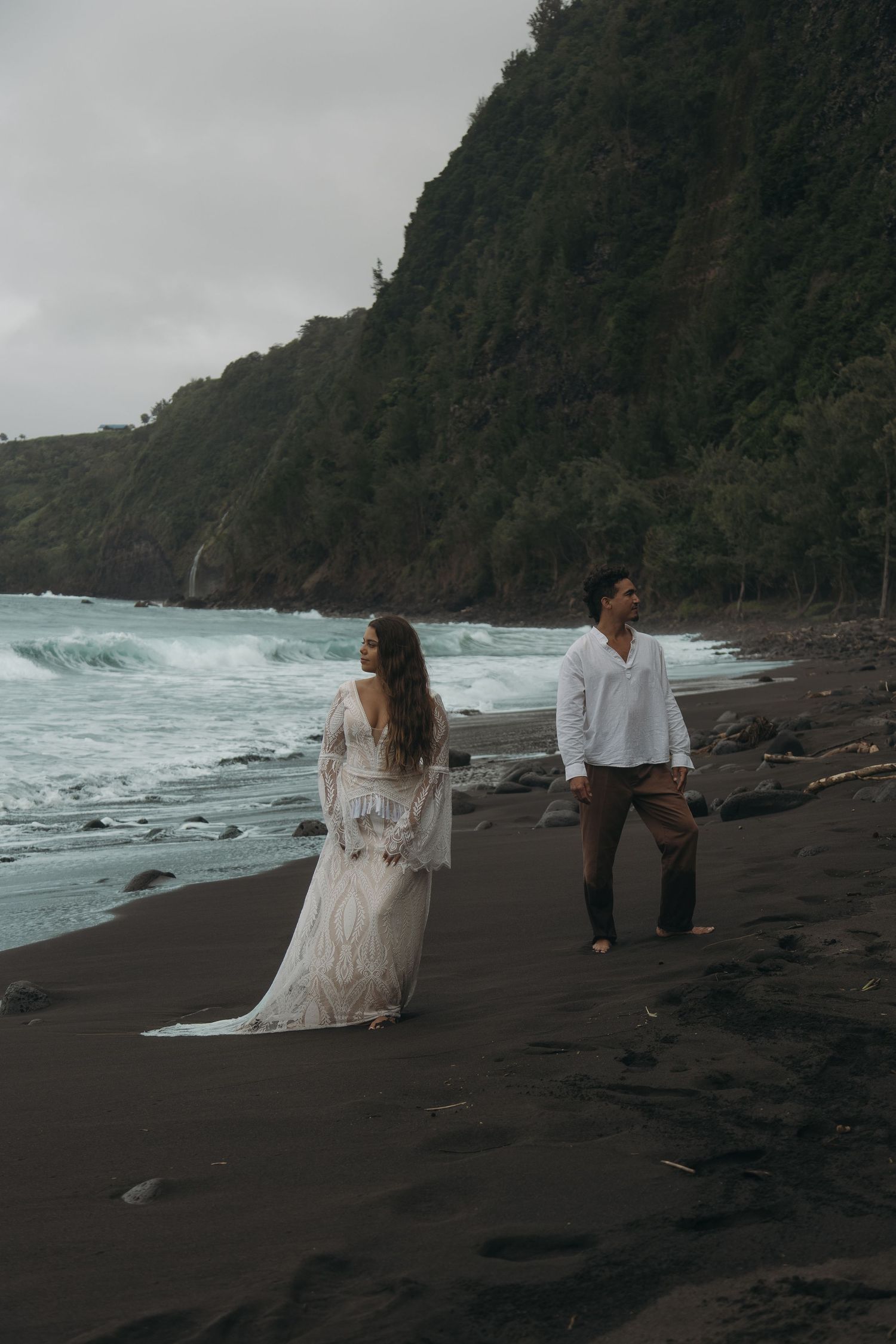 Bride in flowing white lace gown and groom walk along black sand beach with crashing waves.