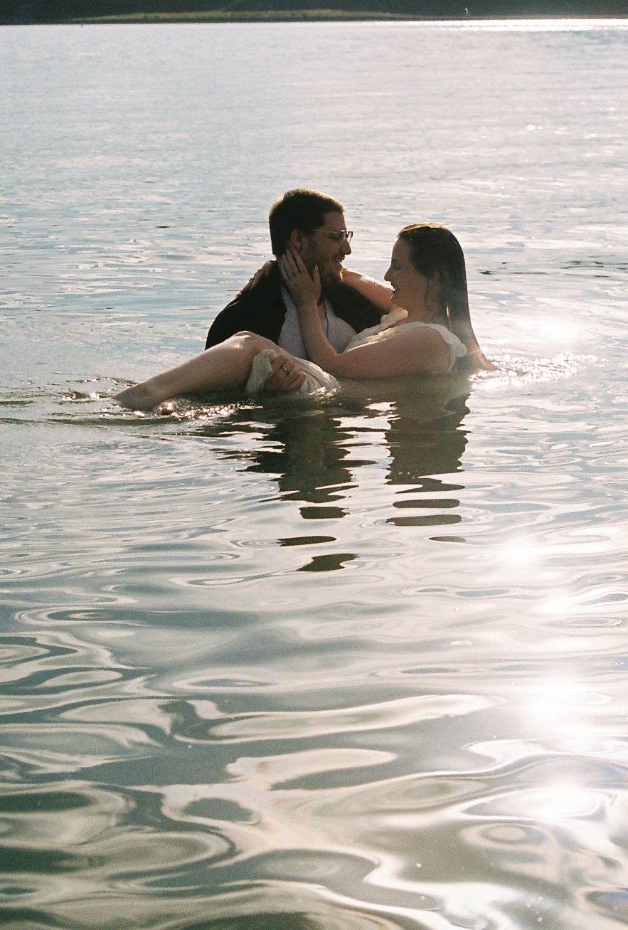 Couple sitting together in sunlit lake water, gazing at each other romantically during golden hour.