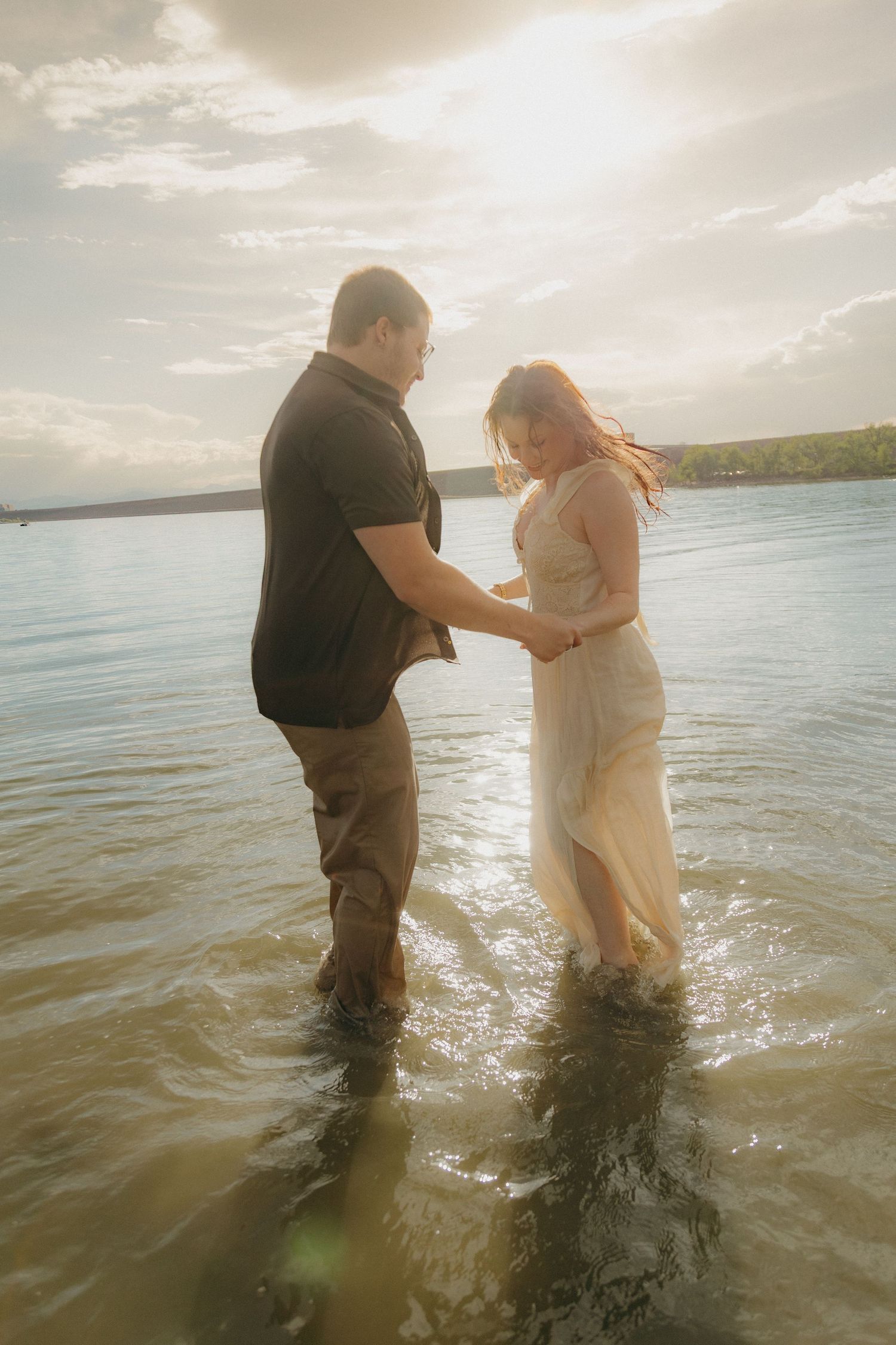 Couple holding hands and standing in shallow lake water at golden hour, woman in white dress.
