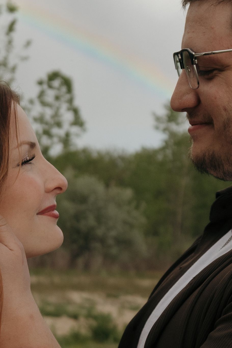 Couple gazing at each other with a rainbow in the background, surrounded by lush green trees outdoors.