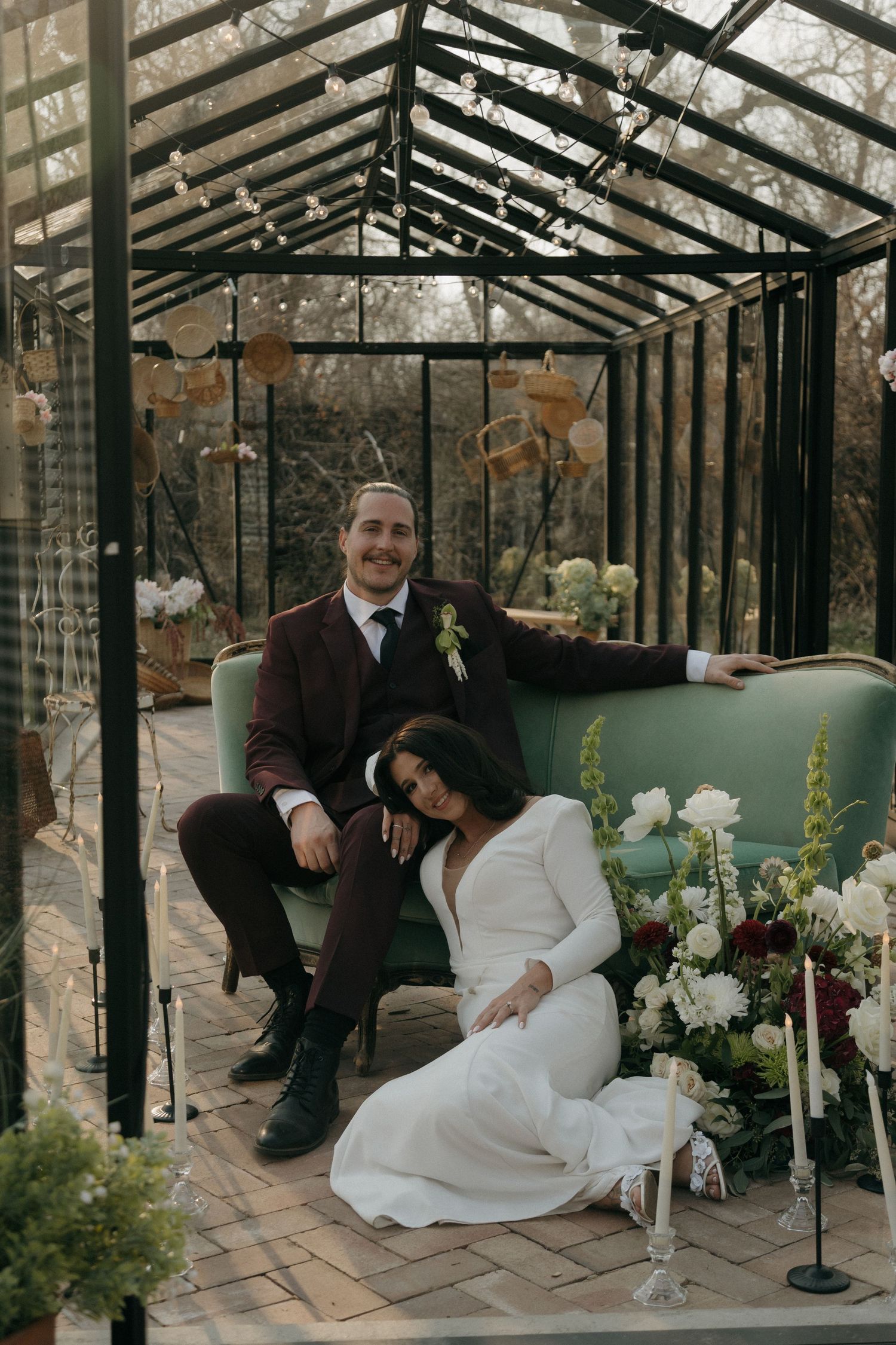 A couple in wedding attire lounges on a stone patio inside a glass greenhouse surrounded by elegant floral arrangements.