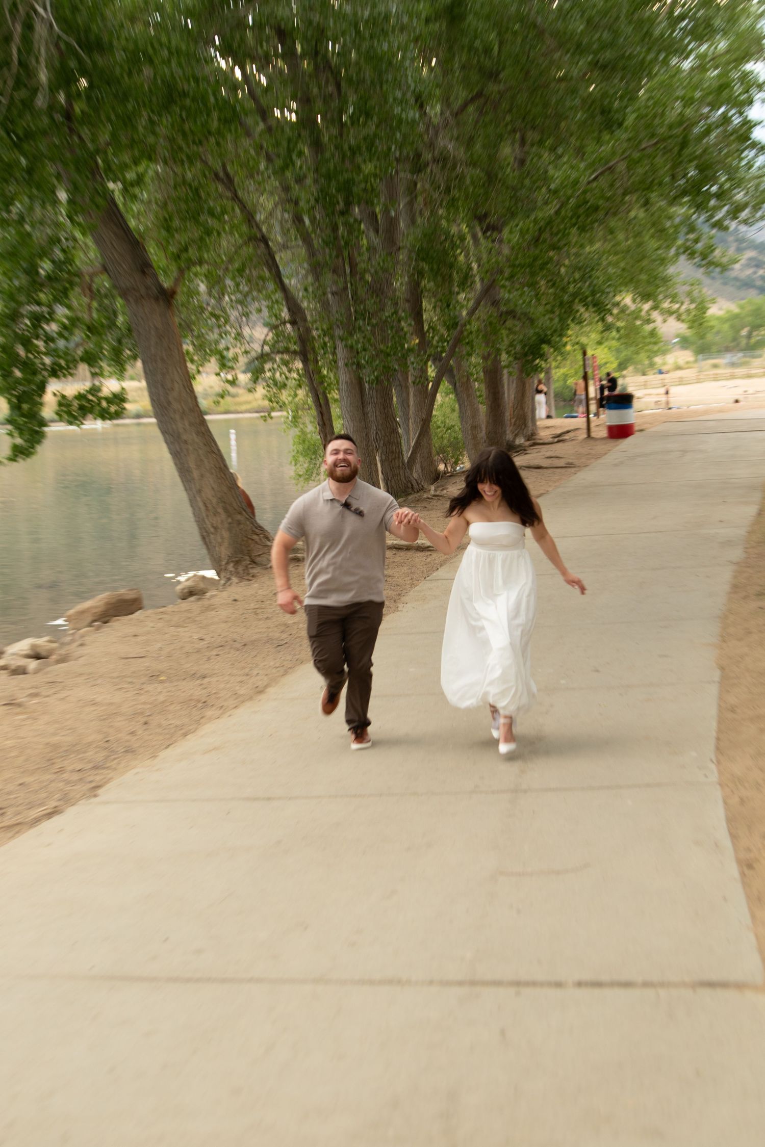 A couple walks hand in hand along a tree-lined path near a lake.