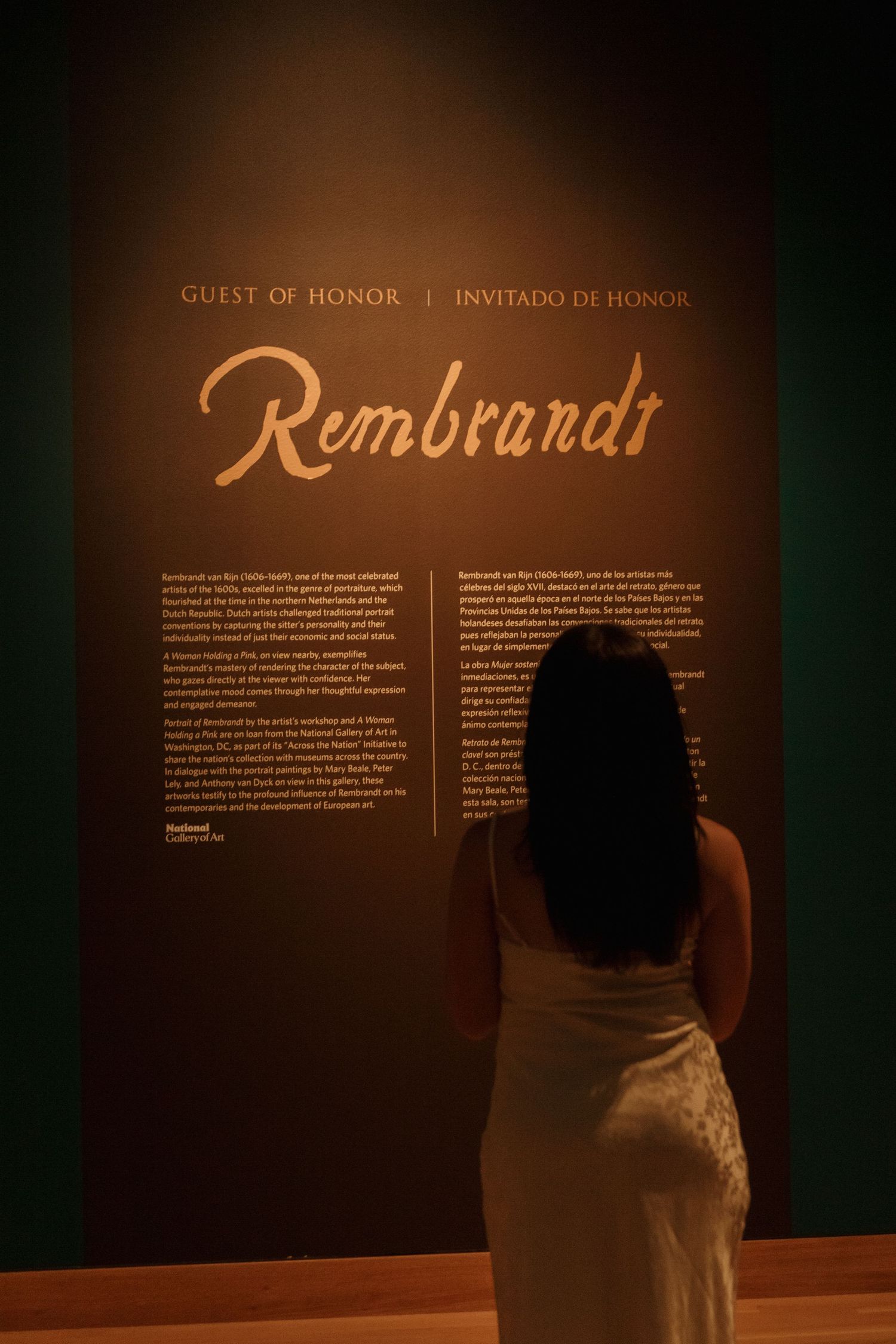 Woman in white dress reading exhibit text about Rembrandt in museum gallery.