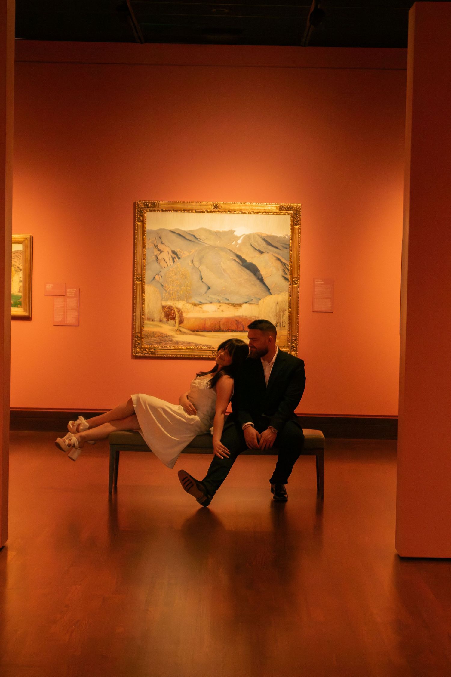 A couple sits together on a bench in an art gallery with framed paintings on coral-colored walls.