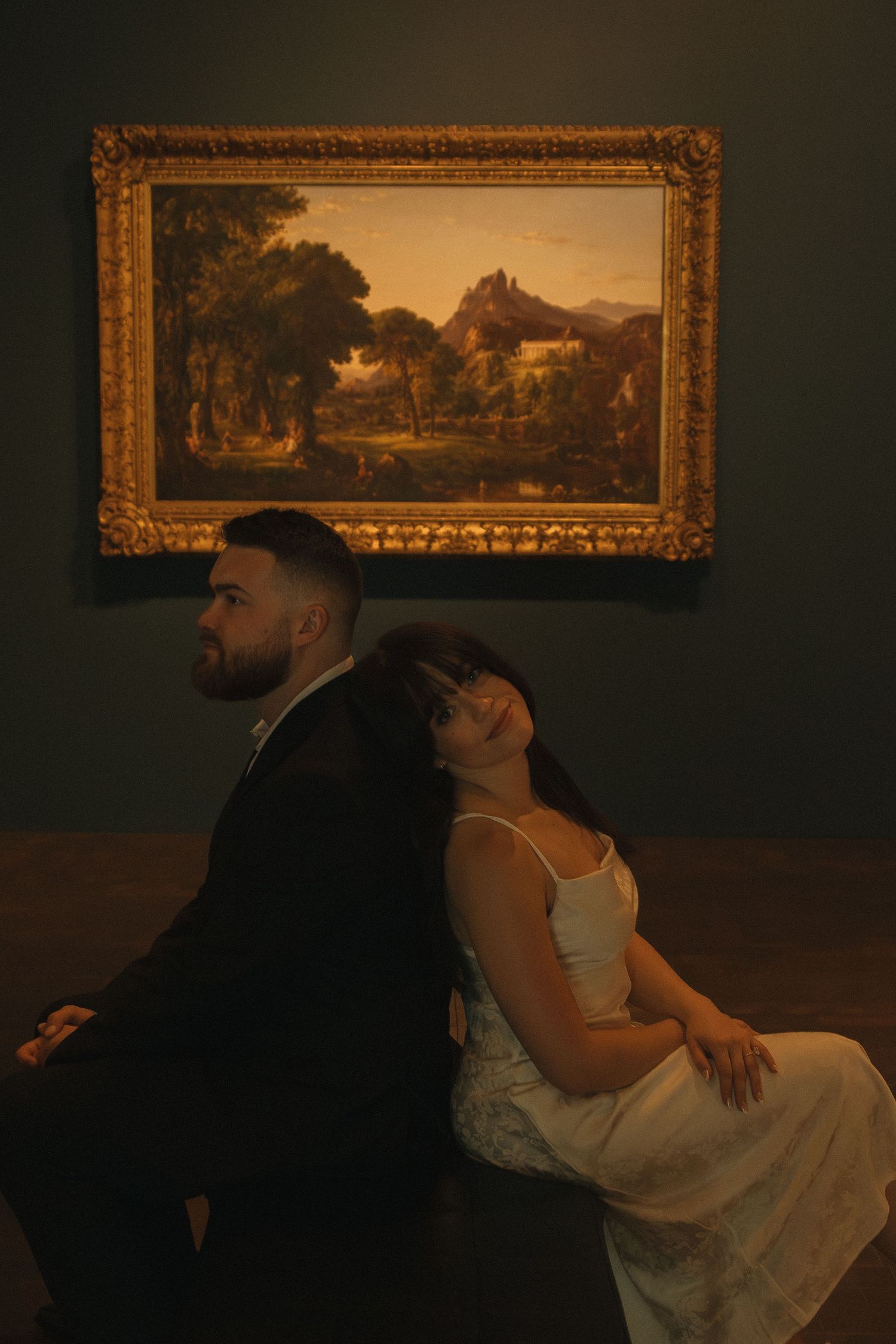 A couple sharing a tender moment in front of ornate gold-framed landscape paintings.