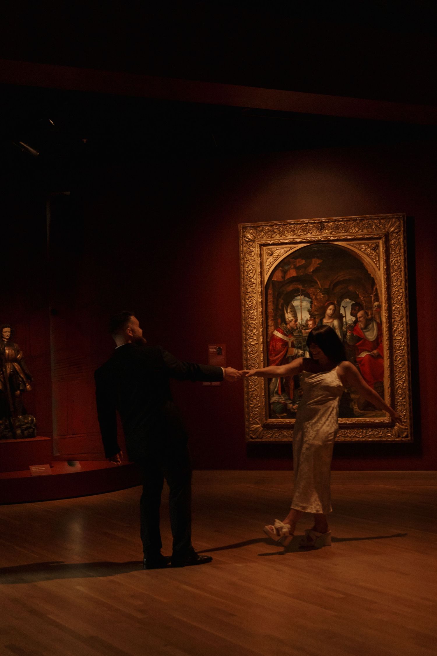 Couples dance in a museum gallery space with classical artwork displayed on red walls.